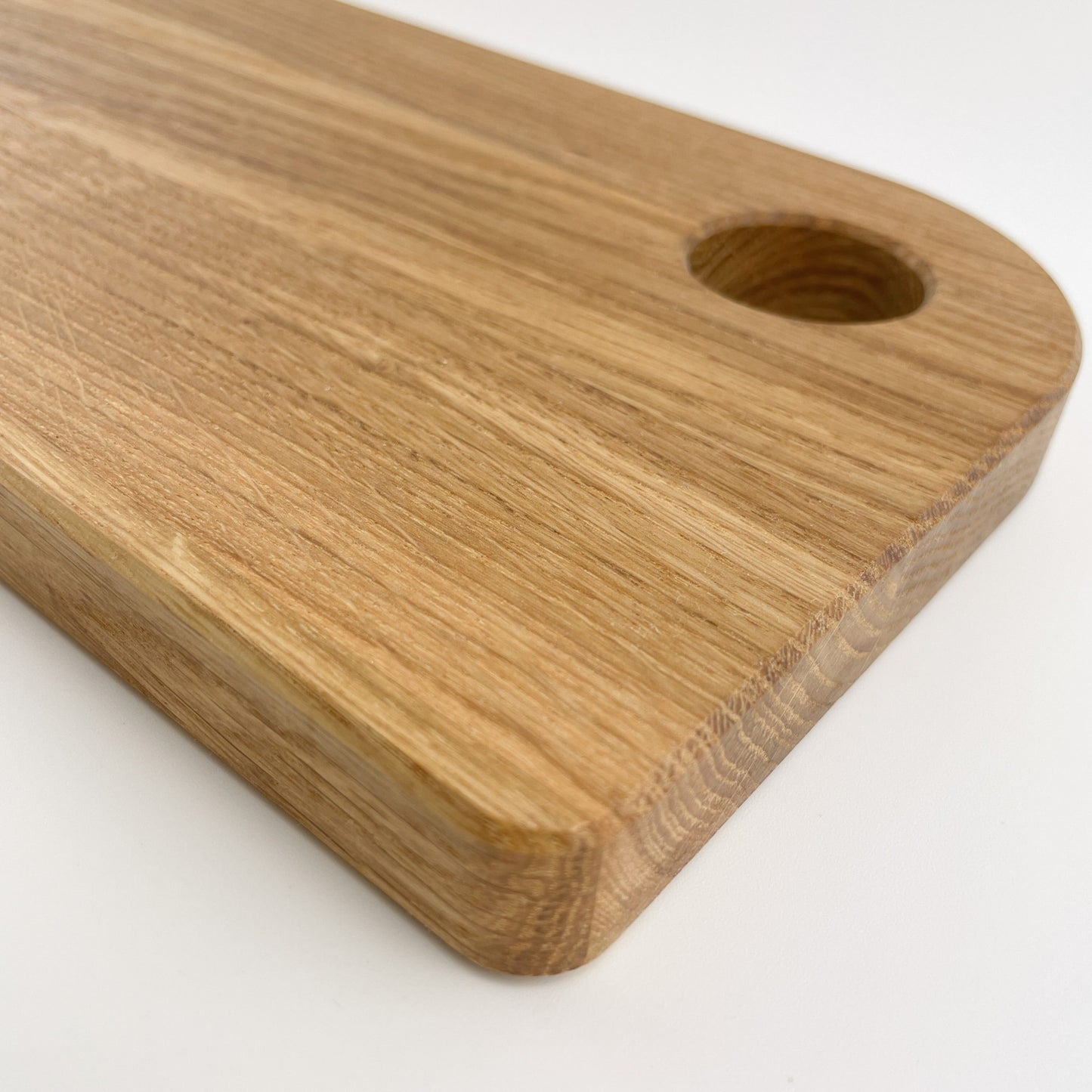 Detailansicht von Holz Schneidebrett - Benno, mit abgerundeten Ecken und einer kleinen kreisförmigen Aussparung am oberen Rand. Das Brett steht auf einer hellen Oberfläche, die einen sanften Kontrast zum warmen Holzton bietet.