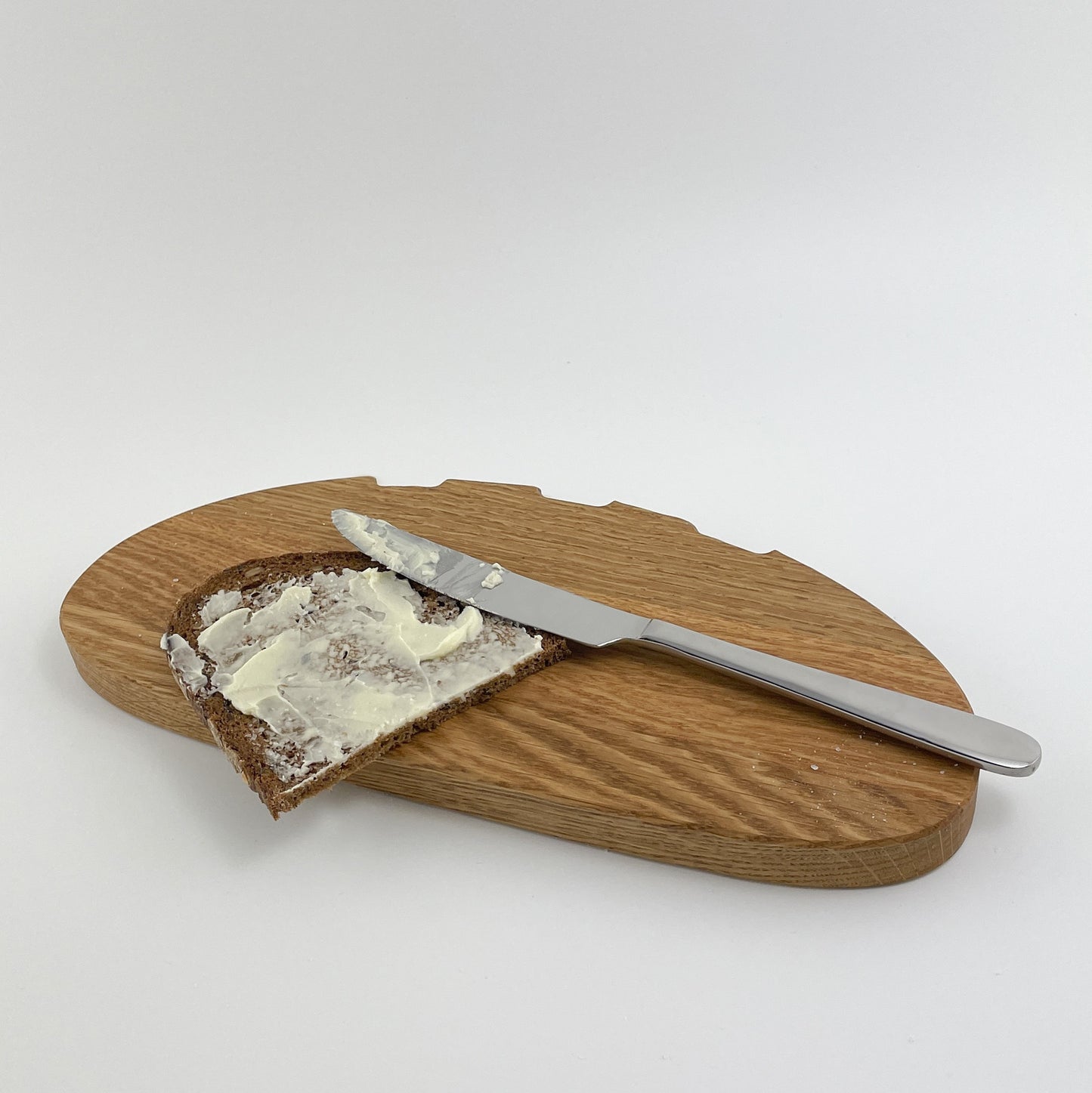 Ein Holz Schneidebrett mit markanter Maserung, genannt Bernd auf dem eine Scheibe Brot liegt. Neben dem Brot befindet sich ein Messer mit silberner Klinge und mattem Griff. Die Szene vermittelt eine alltägliche Küchensituation.