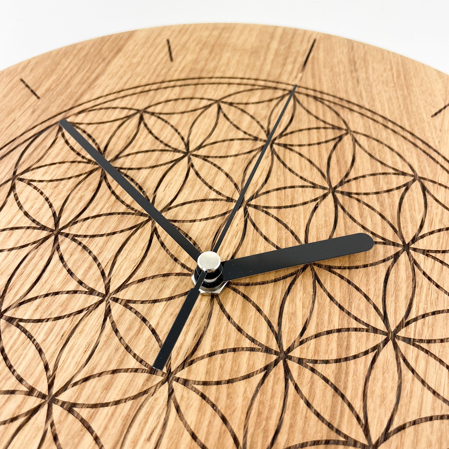 Detailaufnahme von links oben einer Runden Wanduhr aus Massivem Eichenholz (Maserung verläuft vertikal/ aufrecht) mit schwarzen Zeigern vor weißem Hintergrund. Oberfläche wurde mit einem Laser gestaltet und zeigt Die Blume des Lebens im Bereich der Zahlen 11 bis 1.