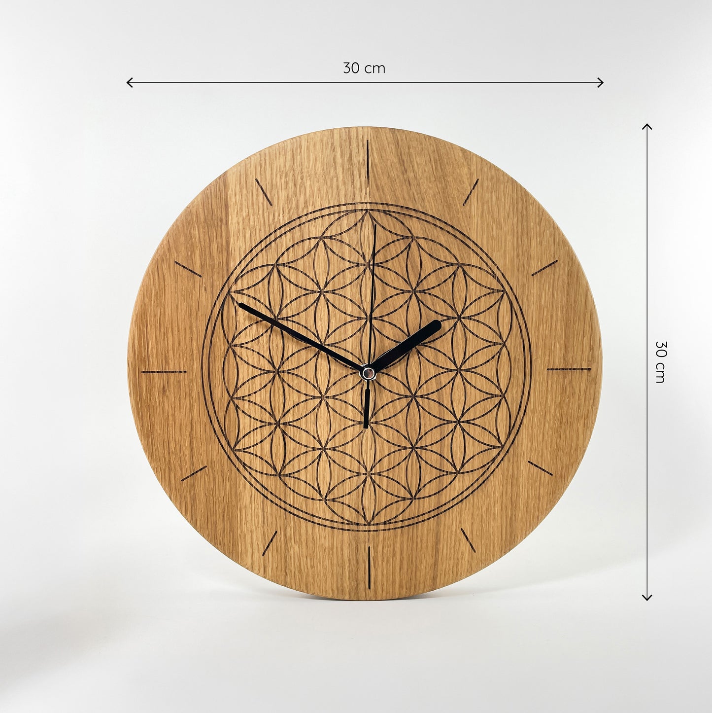Frontalansicht einer Runden Wanduhr aus Massivem Eichenholz (Maserung verläuft vertikal/ aufrecht) mit schwarzen Zeigern vor weißem Hintergrund. Das Zifferblatt ist gelasert und zeigt mittig die Blume des Lebens. Einzelne Linien befinden sich an Stelle der anderen Zahlen. Oberhalb und rechts von der Uhr befinden sich zwei Maßketten, welche ihre Größe angeben – Länge und Höhe sind mit 30cm angegeben.