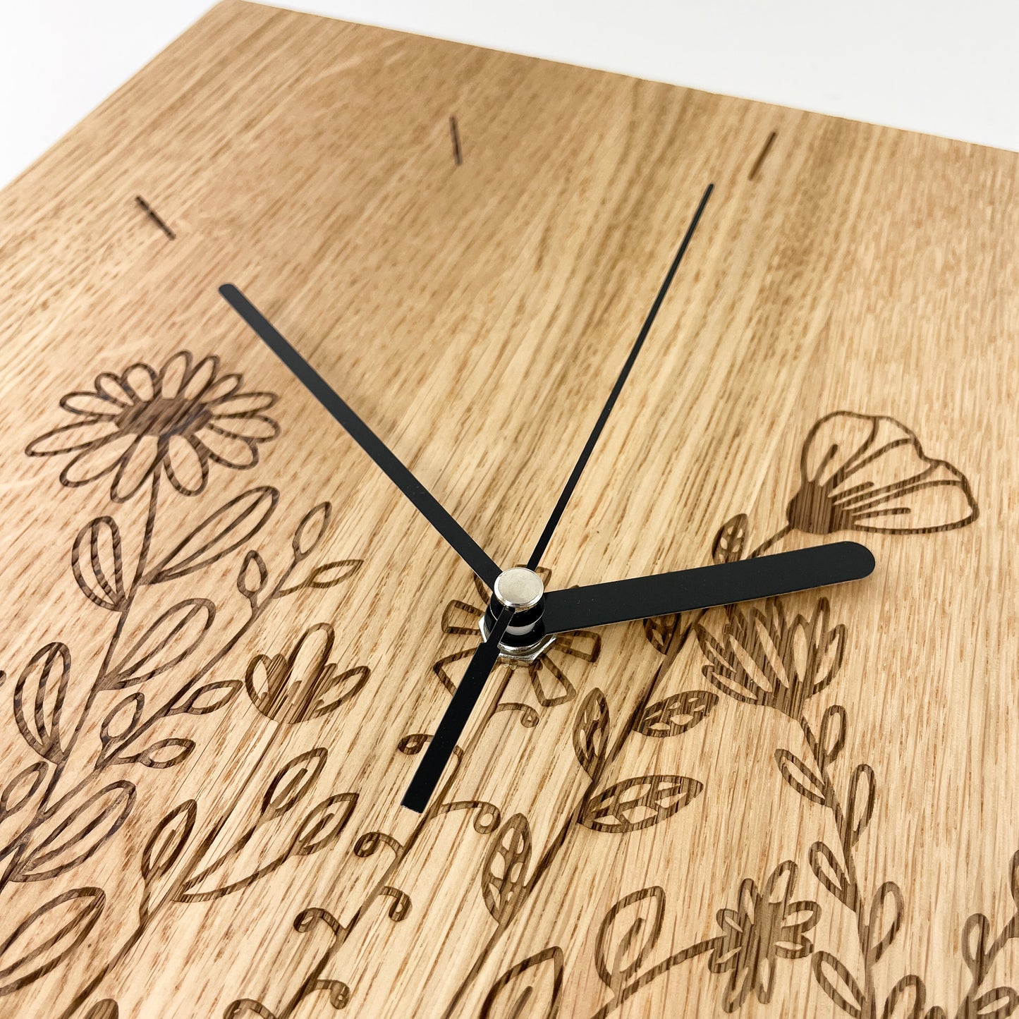 Detailaufnahme von links oben einer quadratischen Wanduhr aus Massivem Eichenholz (Maserung verläuft vertikal/ aufrecht) mit schwarzen Zeigern vor weißem Hintergrund. Die Oberfläche wurde mit einem Laser gestaltet und zeigt einen Teil der Blumenwiese so wie die Striche im Bereich der Zahlen 10-12.