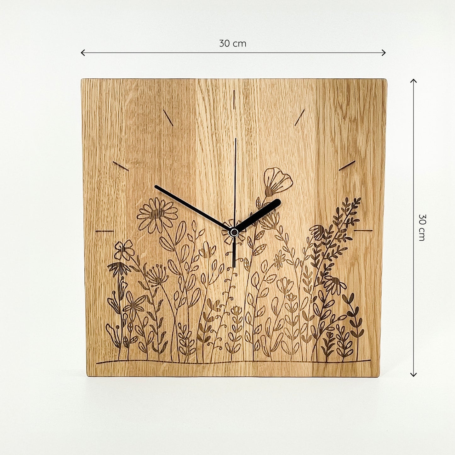 Frontalansicht einer quadratischen Wanduhr aus Massivem Eichenholz (Maserung verläuft vertikal/ aufrecht) mit schwarzen Zeigern vor weißem Hintergrund. Das Zifferblatt ist gelasert und zeigt im unteren Bereich eine Blumenwiese im Bereich der Zahlen 4- 8, Einzelne Linien befinden sich an Stelle der Zahlen 9-3, im Kreisbogen. Oberhalb und rechts von der Uhr befinden sich zwei Maßketten, welche ihre Größe angeben – Länge und Höhe sind mit 30cm angegeben.