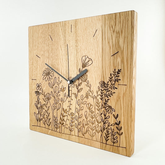 Ansicht einer quadratischen Wanduhr aus Massivem Eichenholz (Maserung verläuft vertikal/ aufrecht) mit schwarzen Zeigern vor weißem Hintergrund. Das Zifferblatt ist gelasert und zeigt im unteren Bereich eine Blumenwiese im Bereich der Zahlen 4- 8, Einzelne Linien befinden sich an Stelle der Zahlen 9-3, im Kreisbogen.