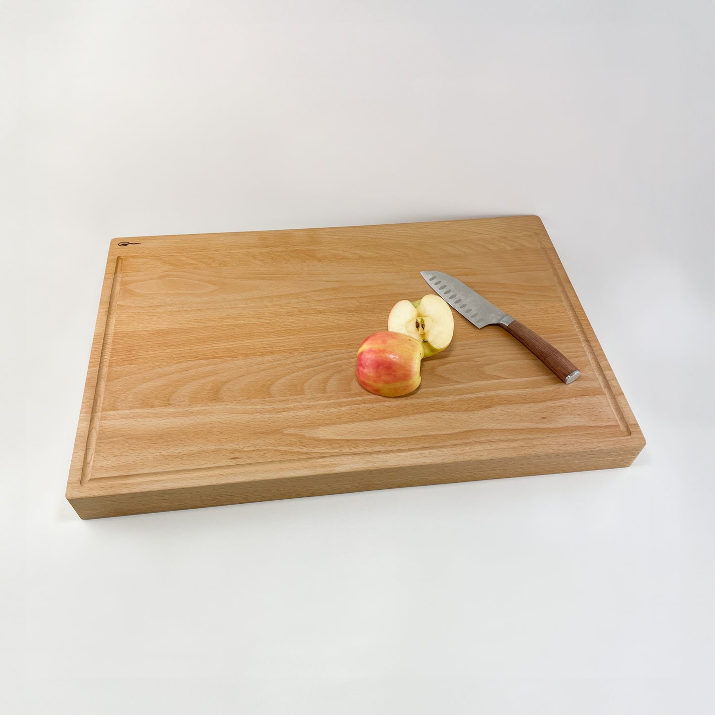Ein Schneidebrett aus Holz, genannt Karl, liegt auf einer weißen Oberfläche. Auf dem Brett befindet sich ein teilweise geschnittener Apfel neben einem Metallmesser mit einem braunen Griff. Die Einfachheit der Szene betont das Zerschneiden von Obst, was mit Kochen oder Lebensmittelvorbereitung in Verbindung stehen kann.