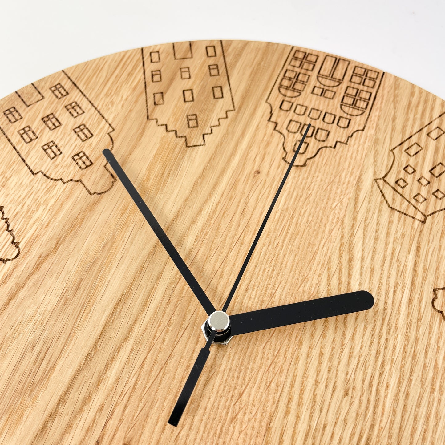 Detailaufnahme von links oben einer Runden Wanduhr aus Massivem Eichenholz (Maserung verläuft vertikal/ aufrecht) mit schwarzen Zeigern vor weißem Hintergrund. Oberfläche wurde mit einem Laser gestaltet und zeigt unterschiedliche Giebelhäuser an Stelle der Zahlen. Darstellung der Giebelhäuser in der Nahaufnahme mit den Häusern 1, 10, 11 und 12.