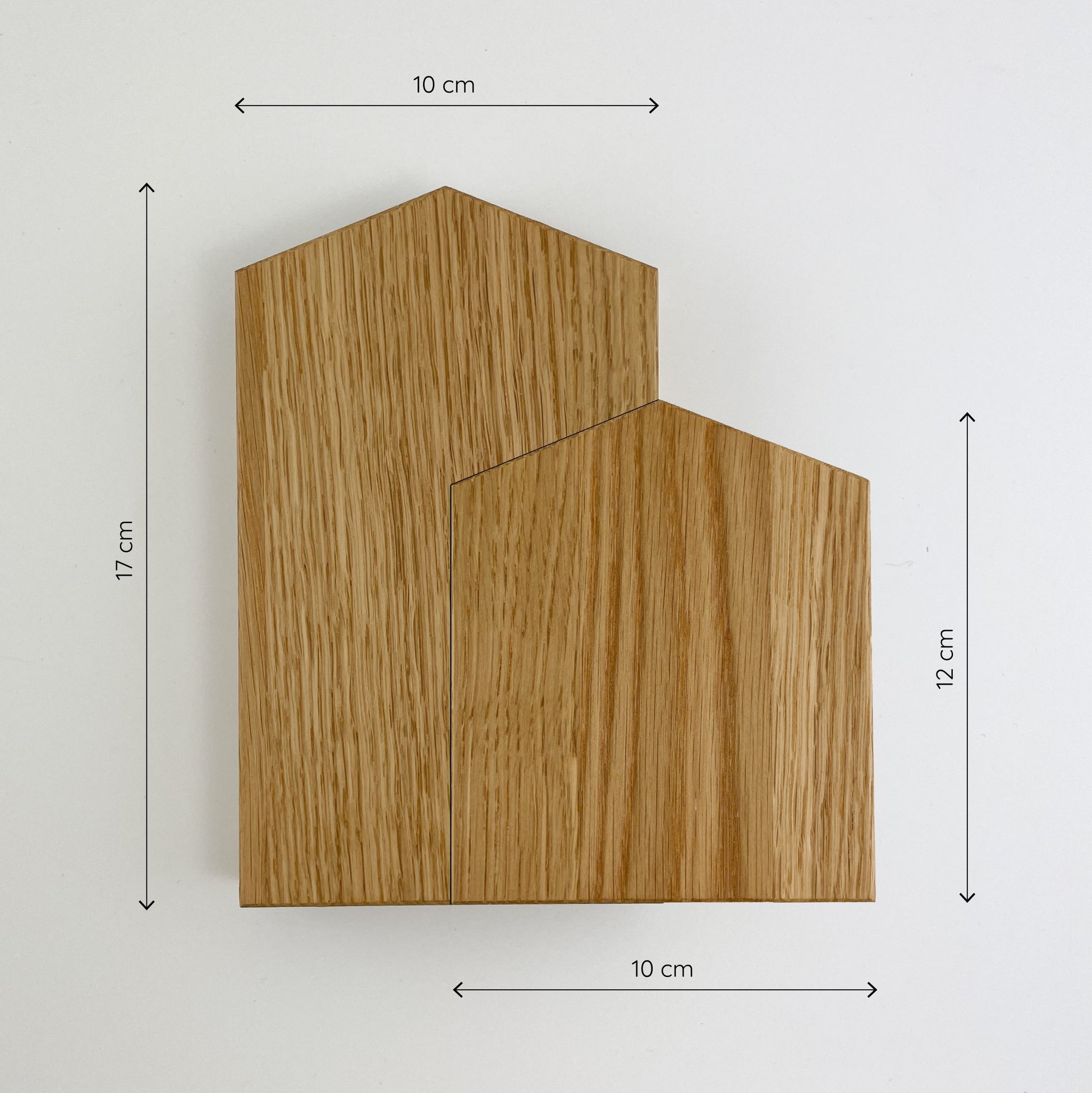 Frontalansicht zweier Deko-Häuser aus (Eichen-) Holz vor weißem Hintergrund. Das linke der beiden Holzhäuser ist etwas größer und die Maßketten links daneben und oben drüber zeigen seine Größe an - es ist 10cm breit und 17cm hoch. Auch das kleinere der beiden Häuser wird von Maßketten definiert und ist ebenfalls 10cm breit, aber nur 12cm hoch. Die Holzmaserung verläuft bei beiden Häusern vertikal.