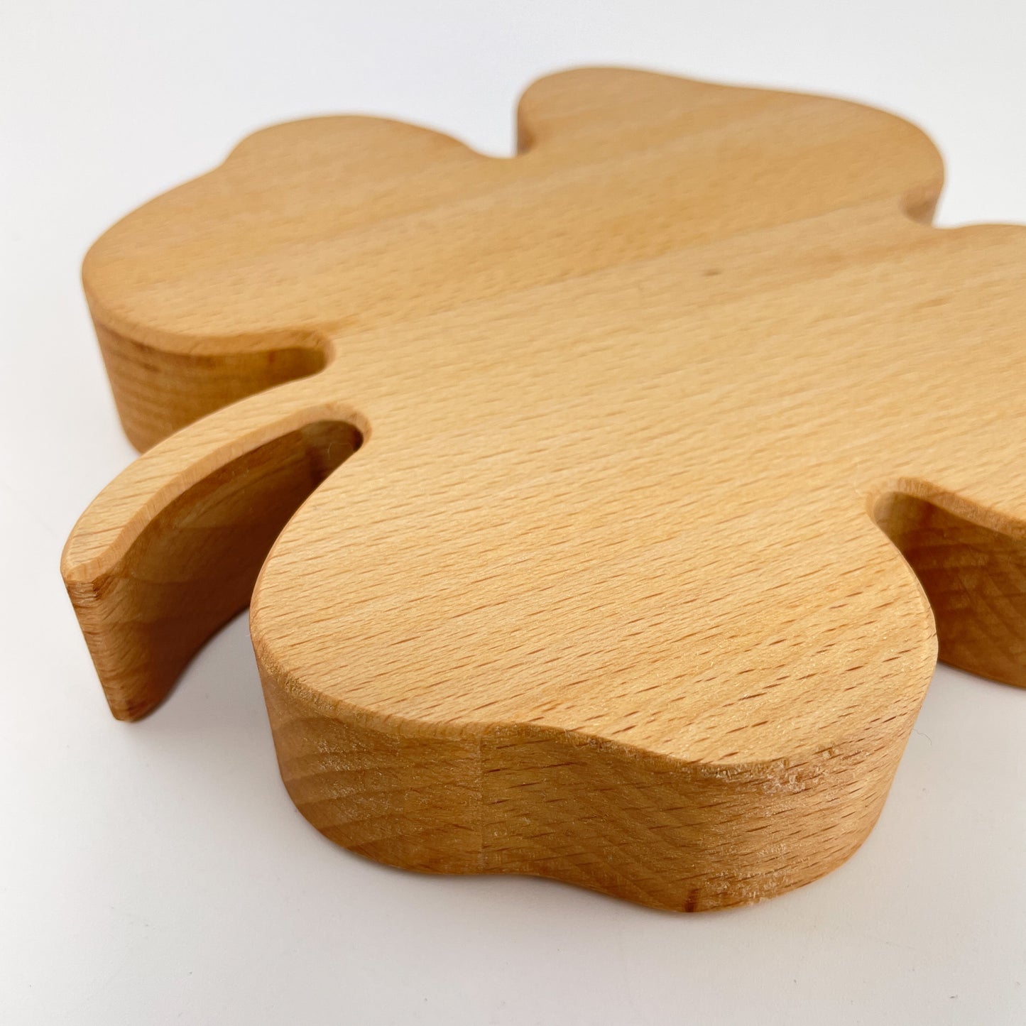 Detailaufnahme eines Holz-Kleeblattes zur Dekoration vor weißem Hintergrund. Der Stiel des vierblättrigen Kleeblattes sowie ein Blatt stehen im Vordergrund, der hintere Teil ist etwas verschwommen. Gut zu sehen ist die abgefaste Kante.