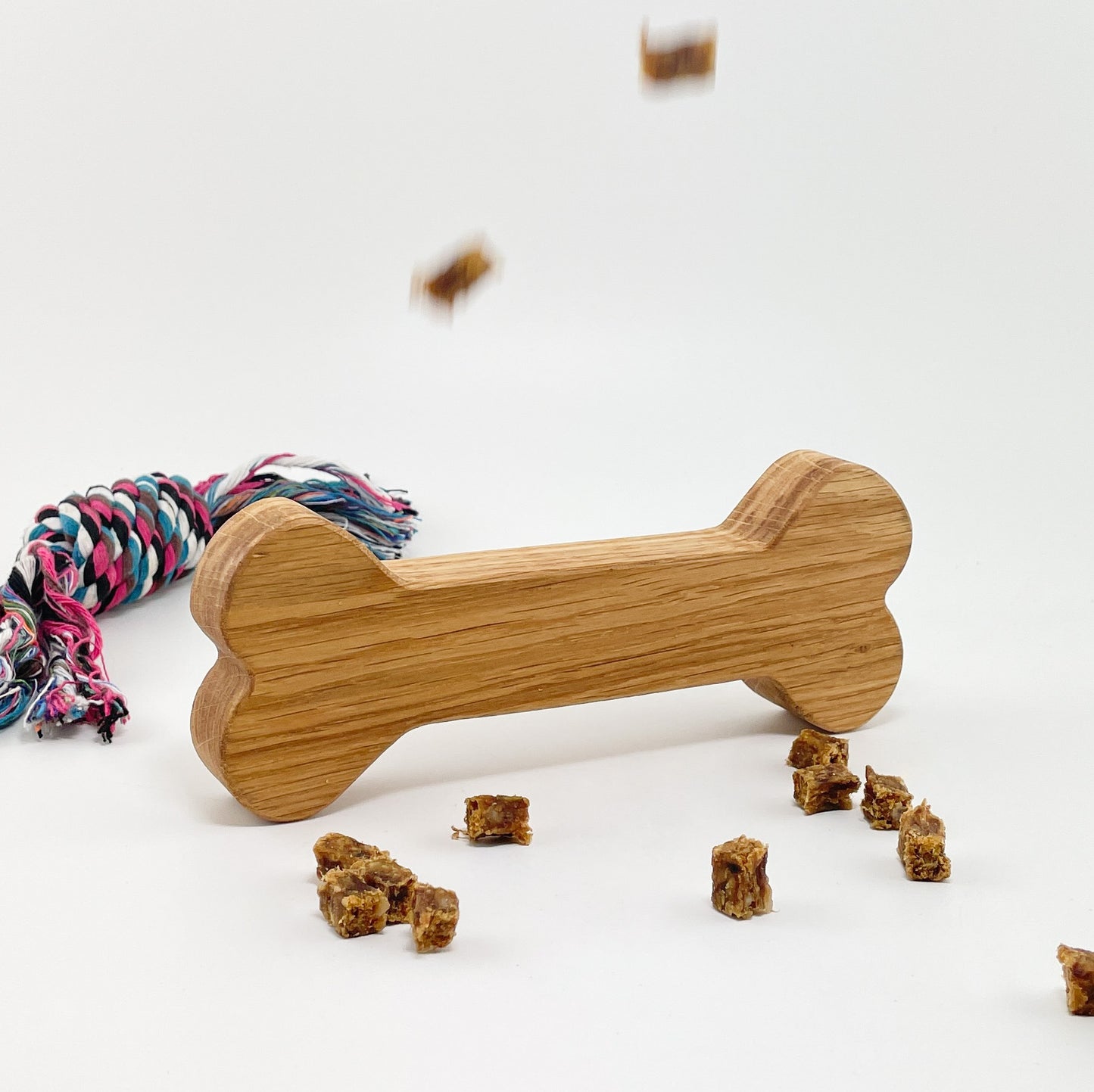 Ein Deko-Knochen aus Holz leicht schräg vor einem weißen Hintergrund aufgestellt. Die Maserung des Eichenholzes verläuft längs auf dem Knochen und im Hintergrund liegt ein Hundespielzeug aus Seil. Vor dem Holzknochen liegen einige Hundeleckerlies verteilt und oben fallen zwei weitere ins Bild.