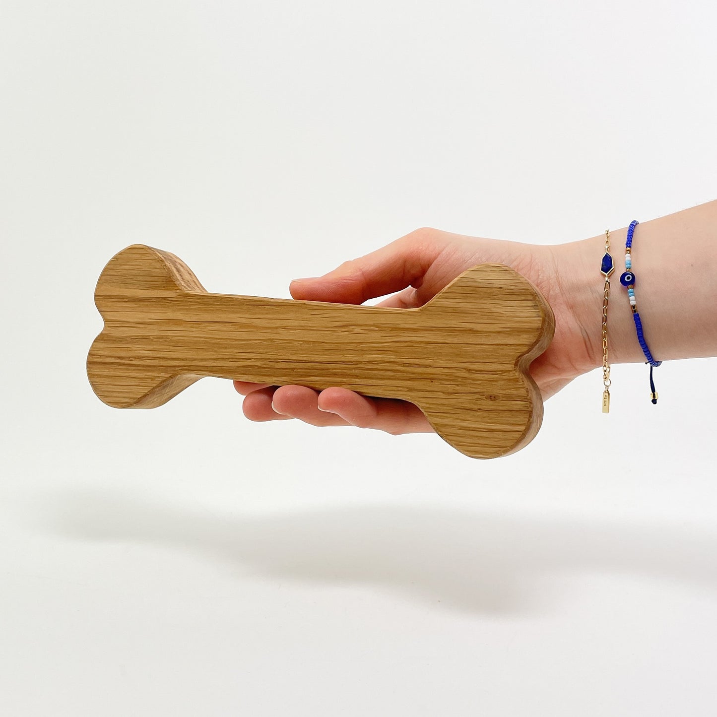 Ein Knochen zur Dekoration aus Eichenholz wird von einer Hand gehalten, die von rechts ins Bild ragt und zwei filigrane blau-goldene Armbänder trägt. Der Hintergrund ist Weiß und die Maserung des Holzes verläuft längs zur Form des Knochens.