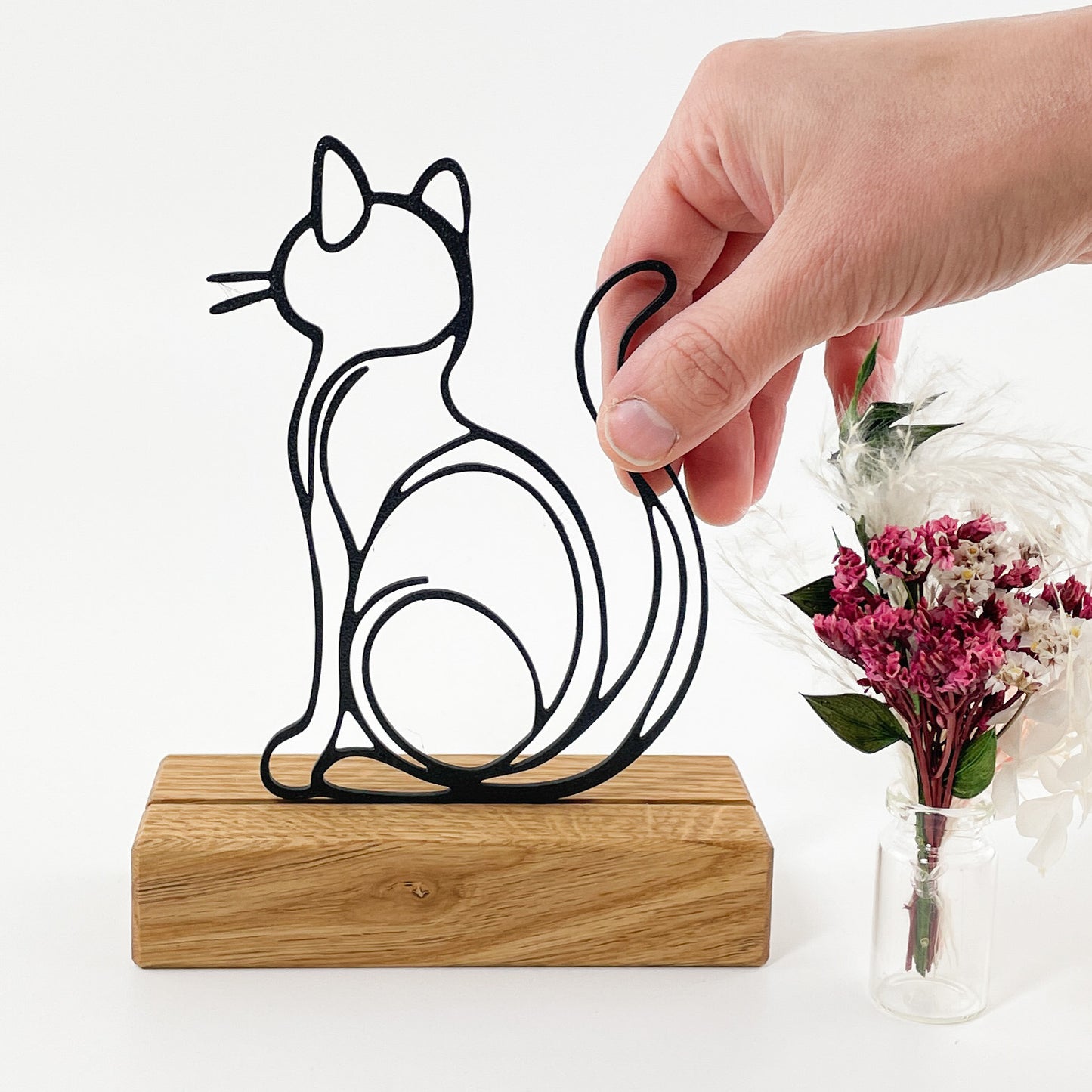 Steckmotiv einer Katze von hinten, sitzend mit aufgerichtetem Schwanz im Stile einer Linienzeichnung, aus schwarzem Kunststoff, welches mit dem 3D-Drucker gefertigt wurde. Die Katze Kira steckt in einem Halter aus Massivholz und links daneben steht ein kleiner Trockenblumenstrauß mit rosa, pinken und weißen Blüten. Von rechts oben ragt eine Hand ins Bild die das Motiv mit 2 Fingern festhält