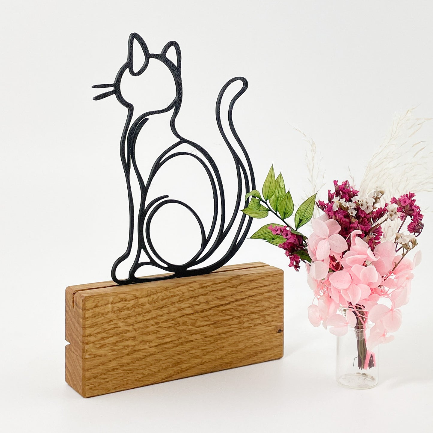 Steckmotiv einer Katze von hinten mit aufgerichtetem Schwanz, sitzend, im Stile einer Linienzeichnung, aus schwarzem Kunststoff, welches mit dem 3D-Drucker gefertigt wurde. Die Katze Kira steckt in einem Halter aus Massivholz und rechts daneben steht ein kleiner Trockenblumenstrauß mit rosa, pinken und weißen Blüten.