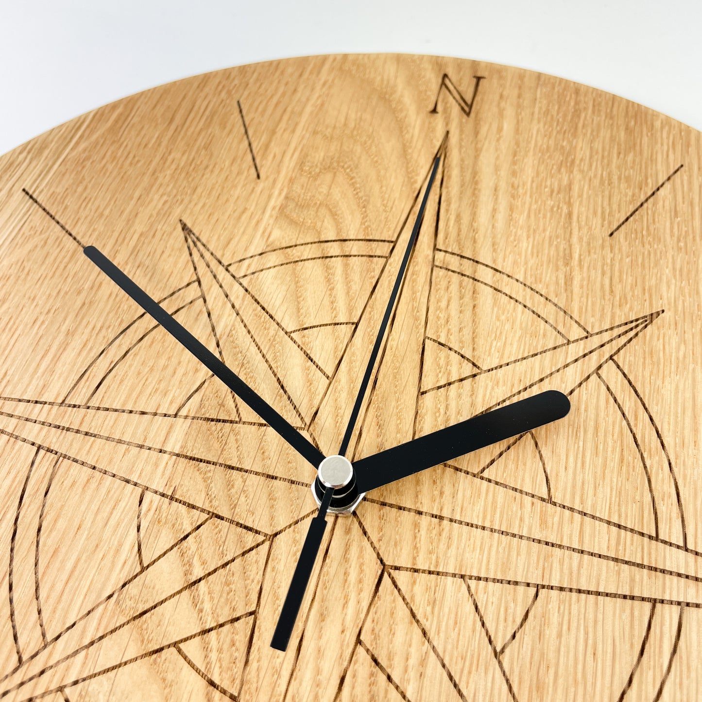 Detailaufnahme von links oben einer Runden Wanduhr aus Massivem Eichenholz (Maserung verläuft vertikal/ aufrecht) mit schwarzen Zeigern vor weißem Hintergrund. Oberfläche wurde mit einem Laser gestaltet und zeigt eine Kompassrose im Bereich der Zahlen 11 bis 1.