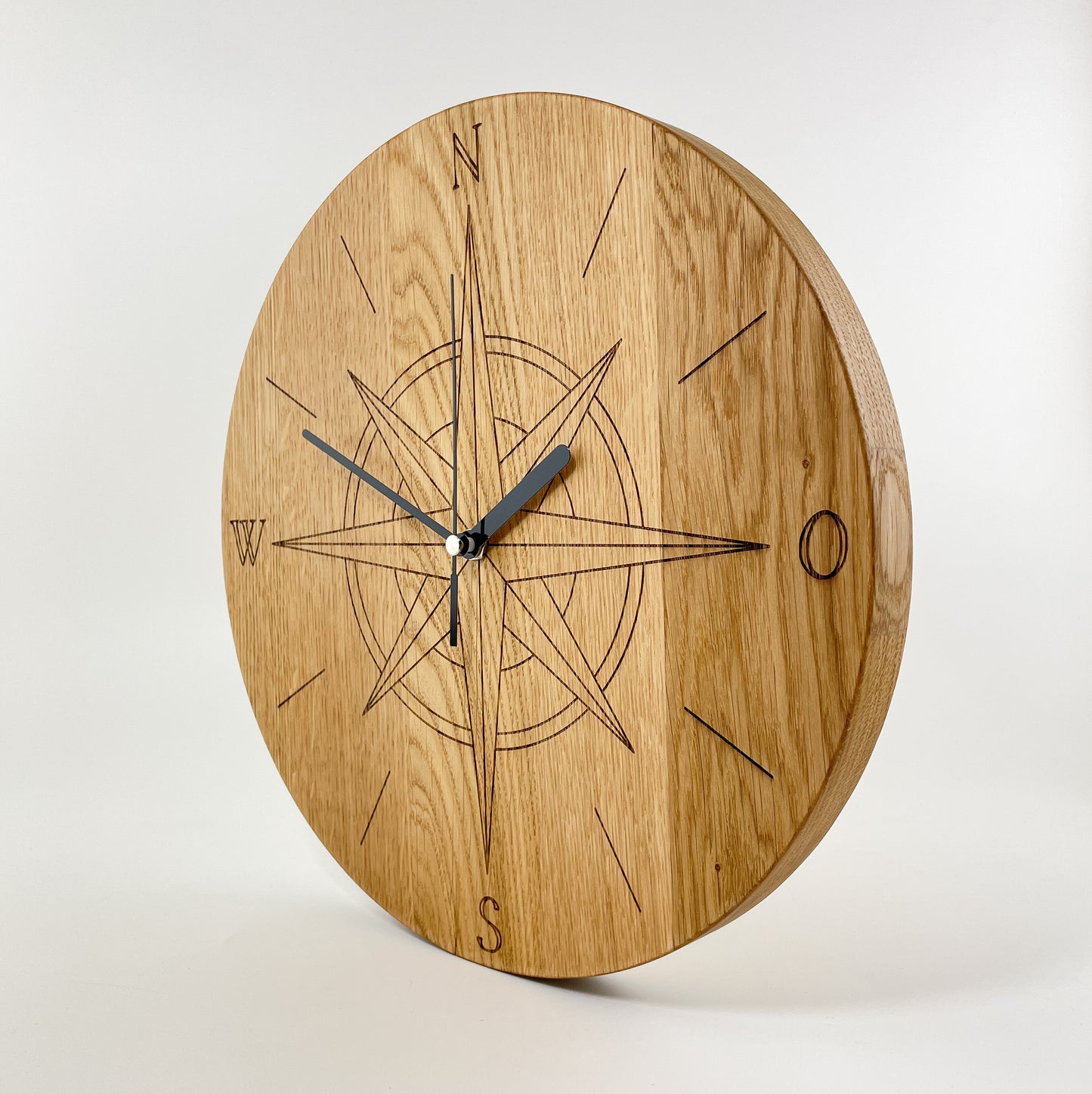 Ansicht einer Runden Wanduhr aus Massivem Eichenholz (Maserung verläuft vertikal/ aufrecht) mit schwarzen Zeigern vor weißem Hintergrund. Das Zifferblatt ist gelasert und zeigt mittig eine Kompassrose, Die Zahlen 3, 6, 9 und 12 sind durch die Himmelsrichtungen ersetzt worden, wobei 12 Norden ist. Einzelne Linien befinden sich an Stelle der anderen Zahlen.