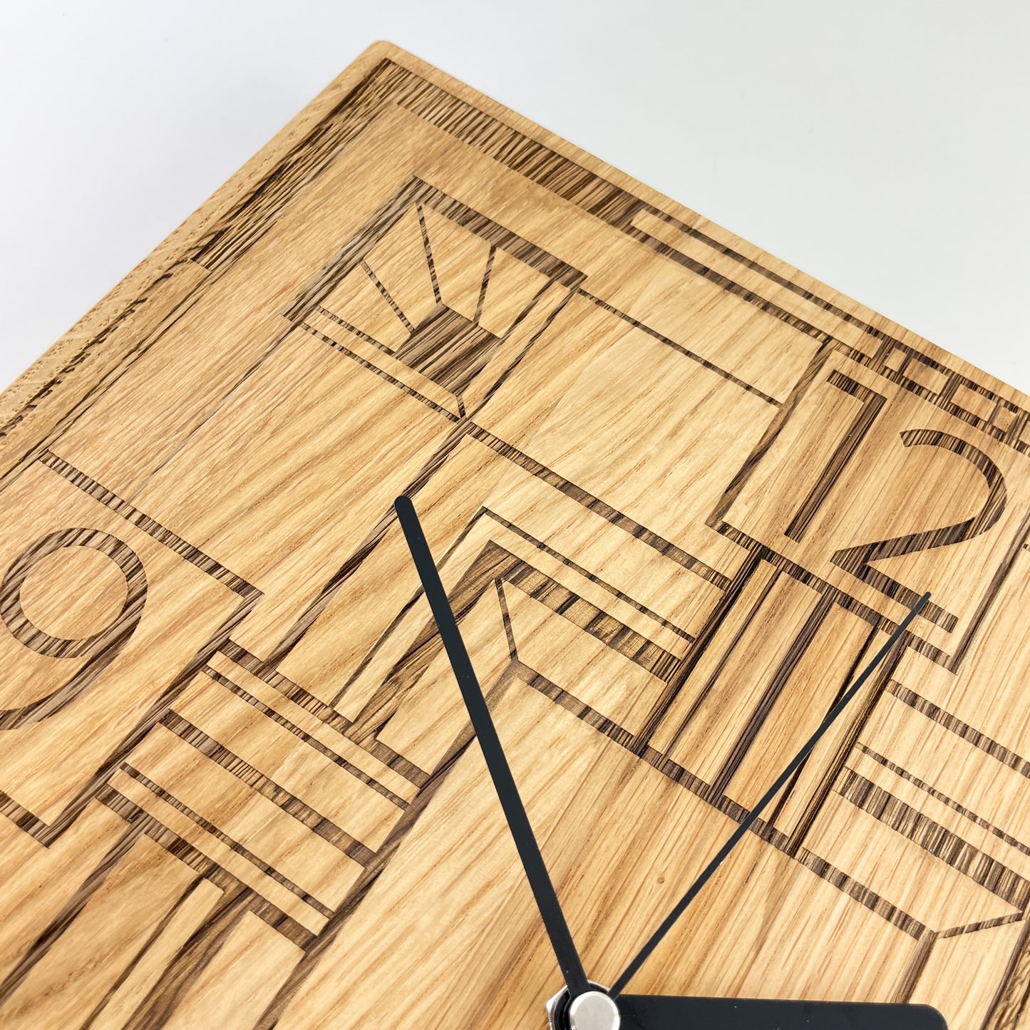 Detail einer quadratischen Wanduhr aus massivem Eichenholz mit senkrechter Maserung und schwarzen Zeigern vor weißem Hintergrund. Das Zifferblatt ist gelasert und zeigt ein geometrisches Art-Déco-Muster mit markanten Linien sowie den Zahlen 12 und 9