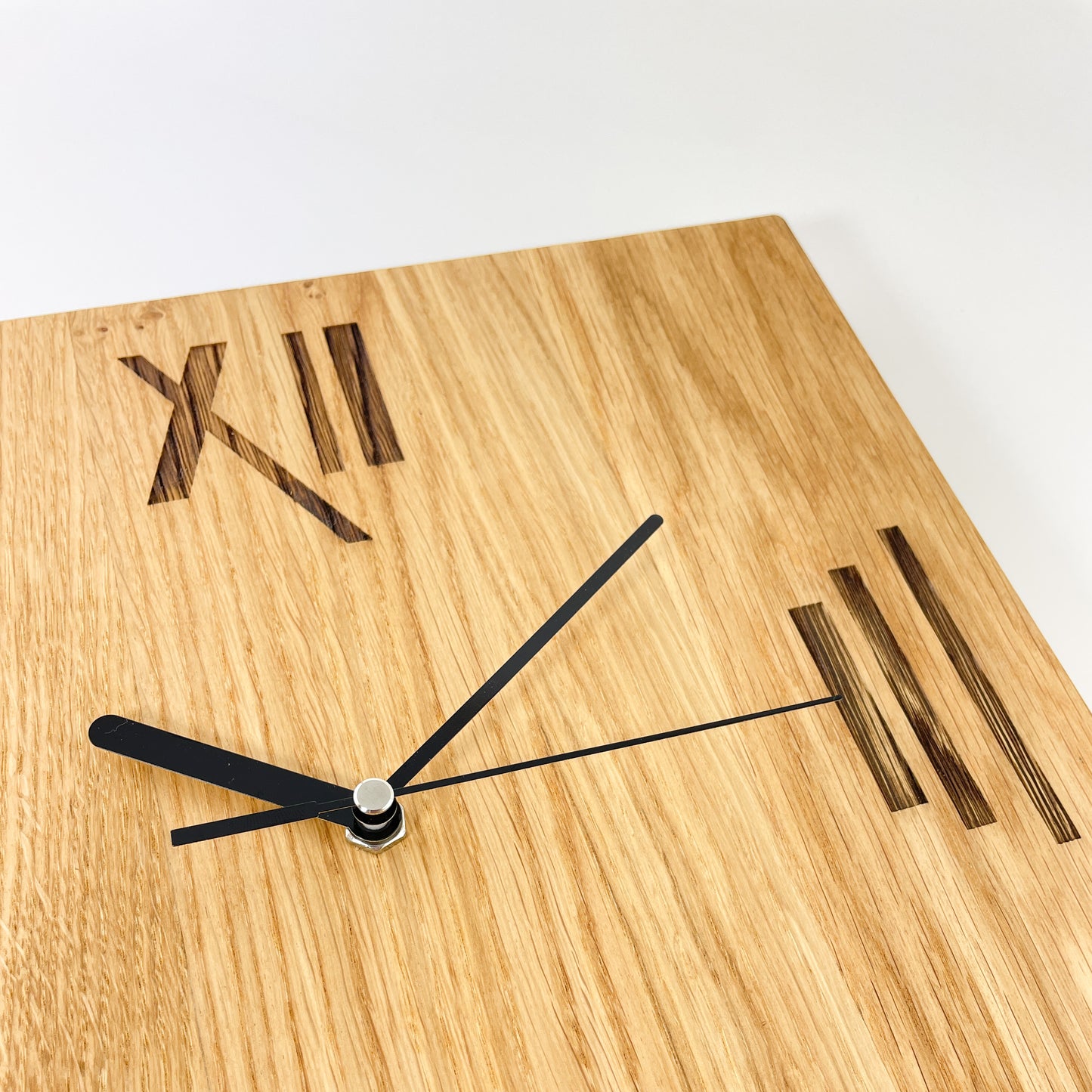 Detailaufnahme der quadratischen Holz-Wanduhr mit senkrechter Maserung und schwarzen Zeigern. Zu sehen sind die römischen Ziffern für die Zahlen 12 und 3, welche grafisch umgesetzt wurden. So besteht die 3 beispielsweise aus drei unterschiedlich langen und dicken Strichen. Die gezeigte Uhrzeit ist 10:10.