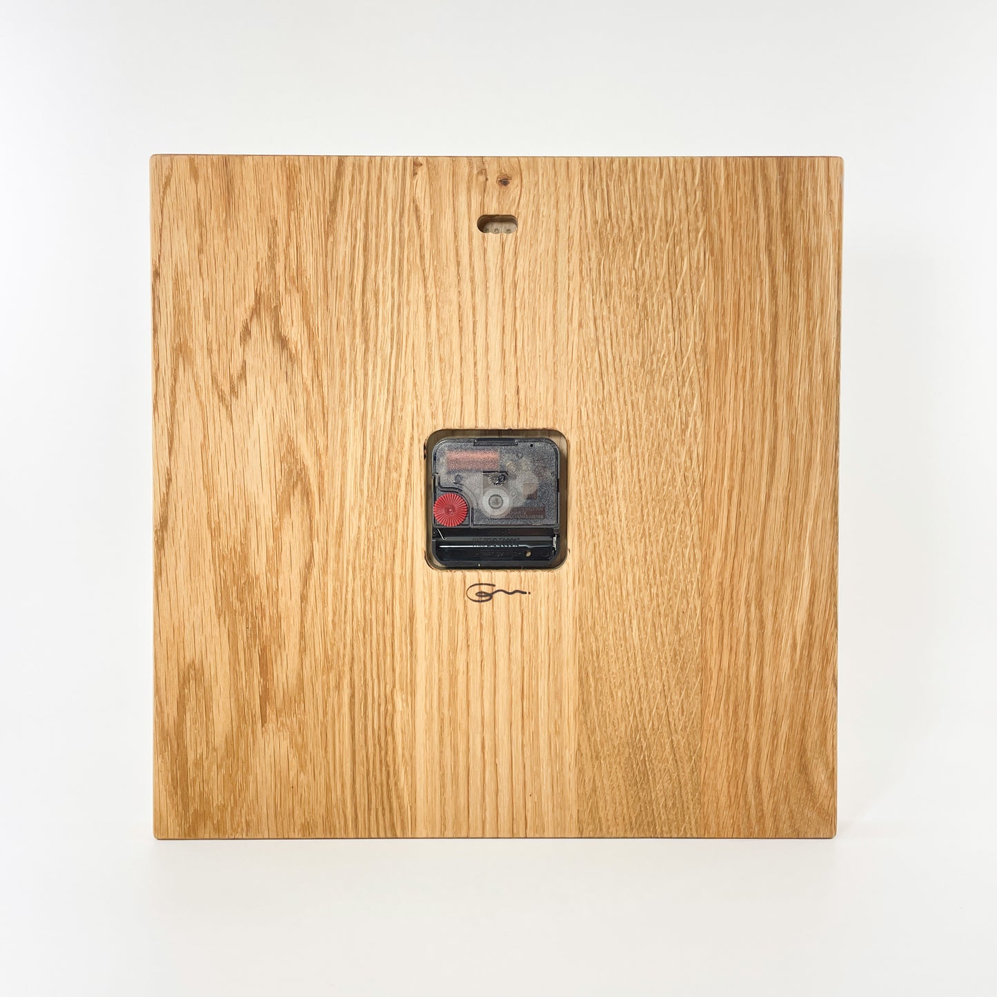 Rückansicht der eckigen Wanduhr aus Holz mit römischen Ziffern auf der Vorderseite. Man sieht mittig platziert das eingelassene Uhrwerk und darunter das dezente Logo der Goertz Möbelmanufaktur Wismar. Oben ist ein Loch für die Aufhängung vorgesehen.