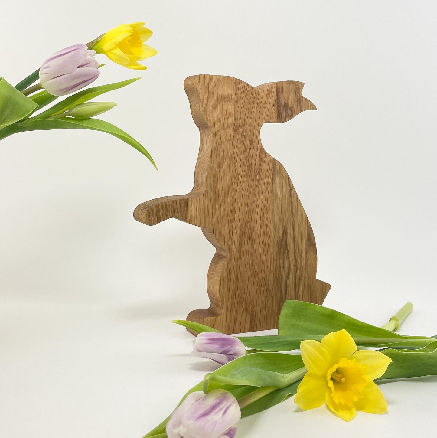 Ein dekorativer Osterhase aus Eichenholz leicht schräg vor weißem Hintergrund. Der Hase macht Männchen und die Maserung des Holzes verläuft senkrecht. Rechts vor dem Hasen liegen zwei lilafarbene Tulpen und eine gelbe Osterglocke. Zudem ragen links im oberen Bereich noch ein paar weitere Blumen ins Bild und es sieht so aus, als würde der Hase daran riechen.