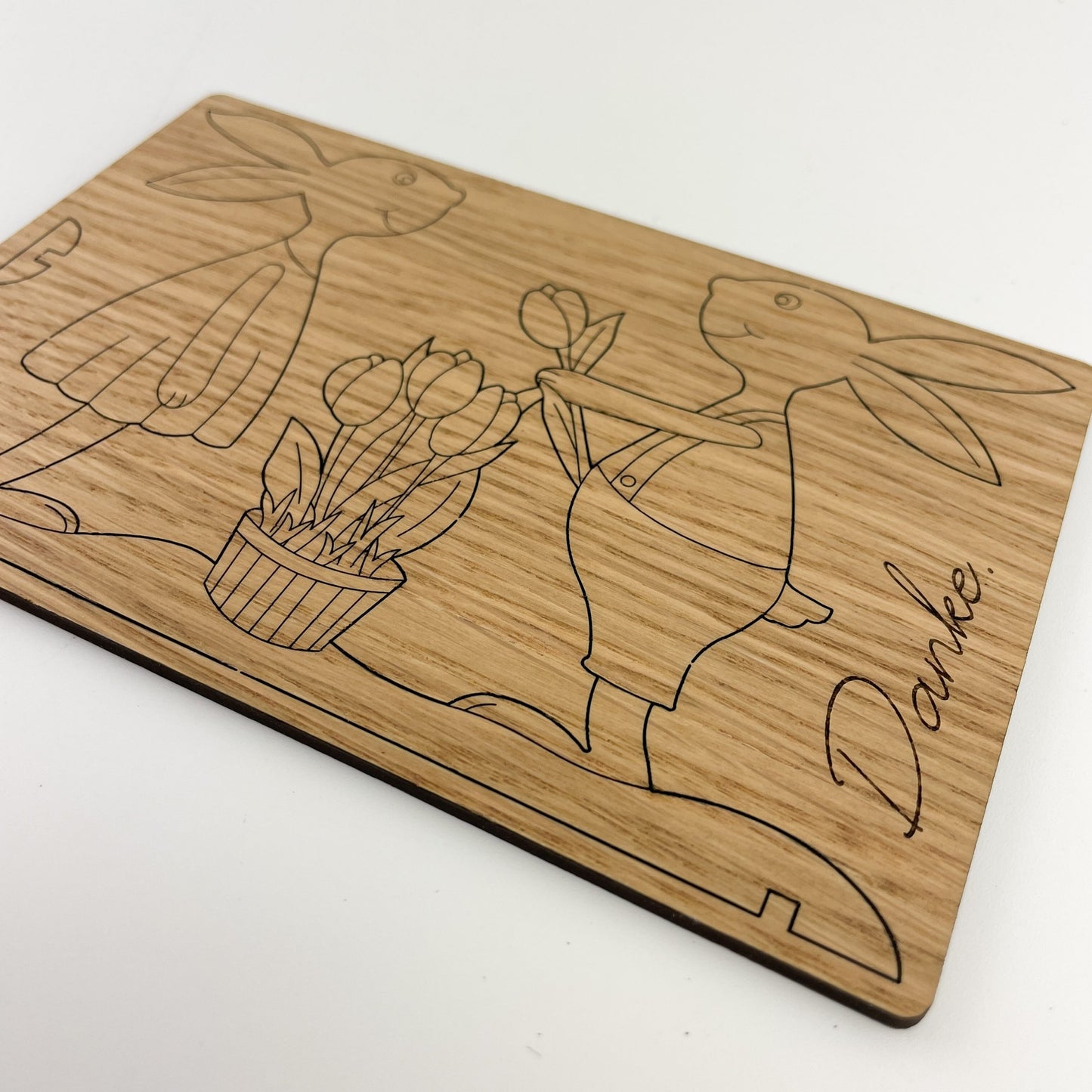 Detailansicht von Holz-Osterkarte mit eingravierten Motiven von zwei Hasen, Papaja & Pepito genannt, die Frühlingsblumen halten. Der linke Hase trägt ein Kleid und hält einen Korb voller Tulpen, während der rechte Hase eine einzelne Blume präsentiert. Unter den Figuren steht das Wort “Danke” in geschwungener Schrift. Die natürliche Maserung des Holzes verleiht der Karte eine warme Textur und betont die handwerkliche Qualität des Gegenstands.