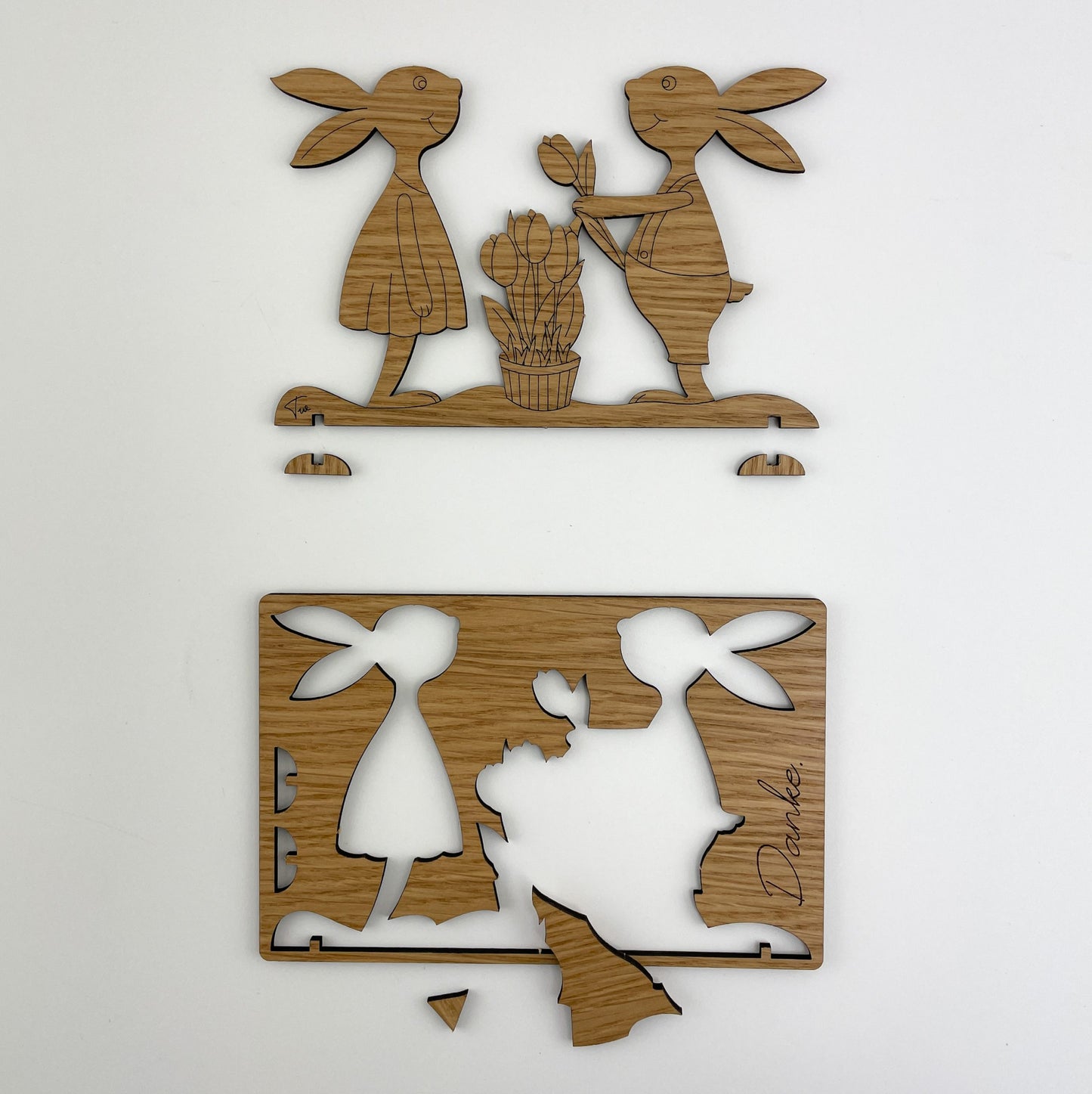 Zwei hölzerne Osterkarten mit ausgeschnittenen Motiven. Die obere Karte zeigt die ausgeschnittenen Kaninchen. Beide sind auf einer Plattform mit dem Schriftzug “Papaja & Pepito” verbunden. Die untere Karte zeigt die Silhouetten von zwei Hasen nebeneinander, wobei ein Teil des Materials innerhalb der Konturen entfernt wurde, um die Formen hervorzuheben.