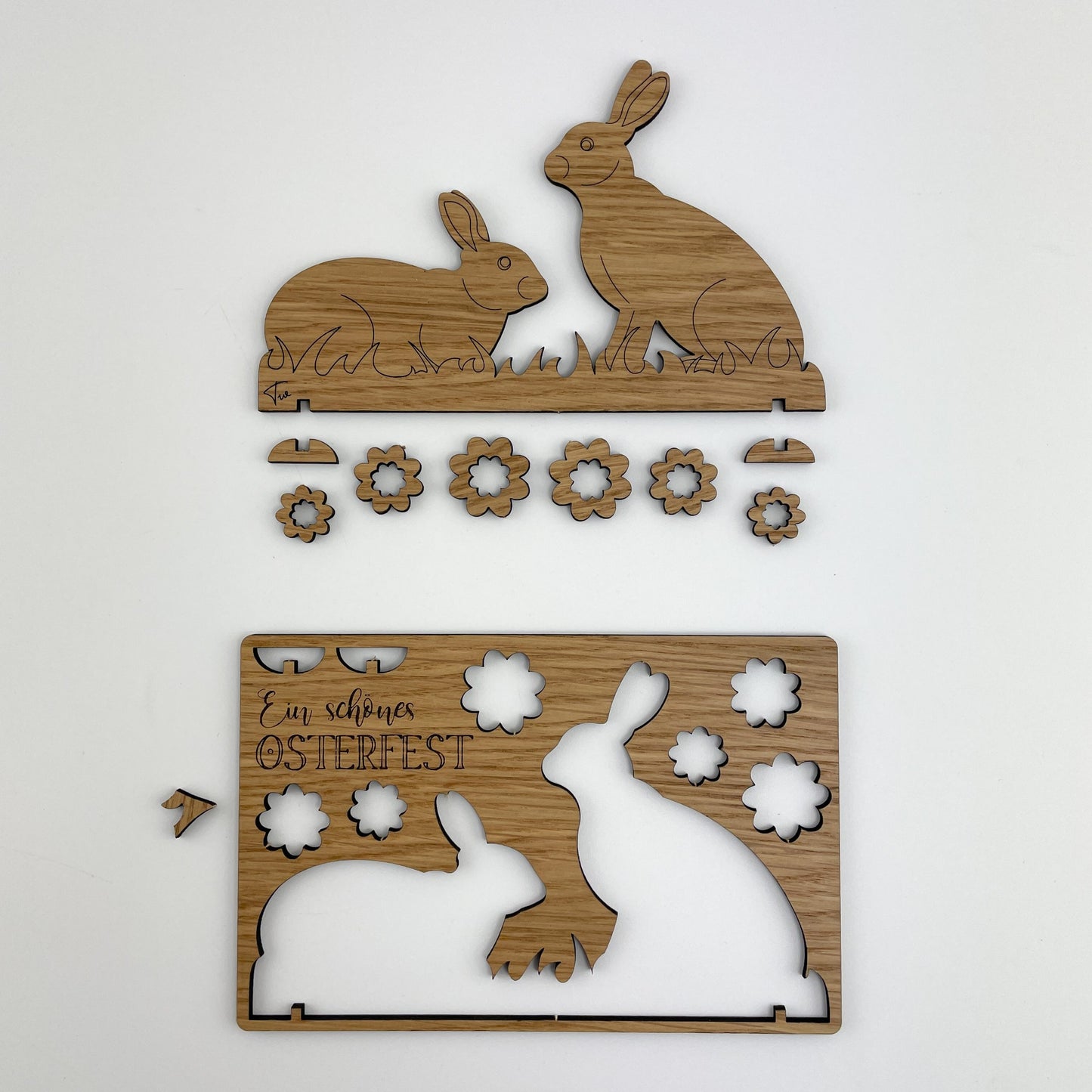 Frontalansicht von Holz-Osterkarte Pia & Pedroist, ein künstlerisch gestaltetes Stück, das aus einer rechteckigen Holztafel besteht. Im oberen Teil der Karte sind zwei Hasen abgebildet, die sich gegenüberstehen. Diese Hasen sind von kleineren Blumen und einem Schmetterling umgeben. Die Figuren sind präzise ausgeschnitten und scheinen abnehmbar zu sein. Neben den Kaninchen gibt es mehrere Blumen und einen Schmetterling, die ebenfalls als herausnehmbare Elemente gestaltet sind.