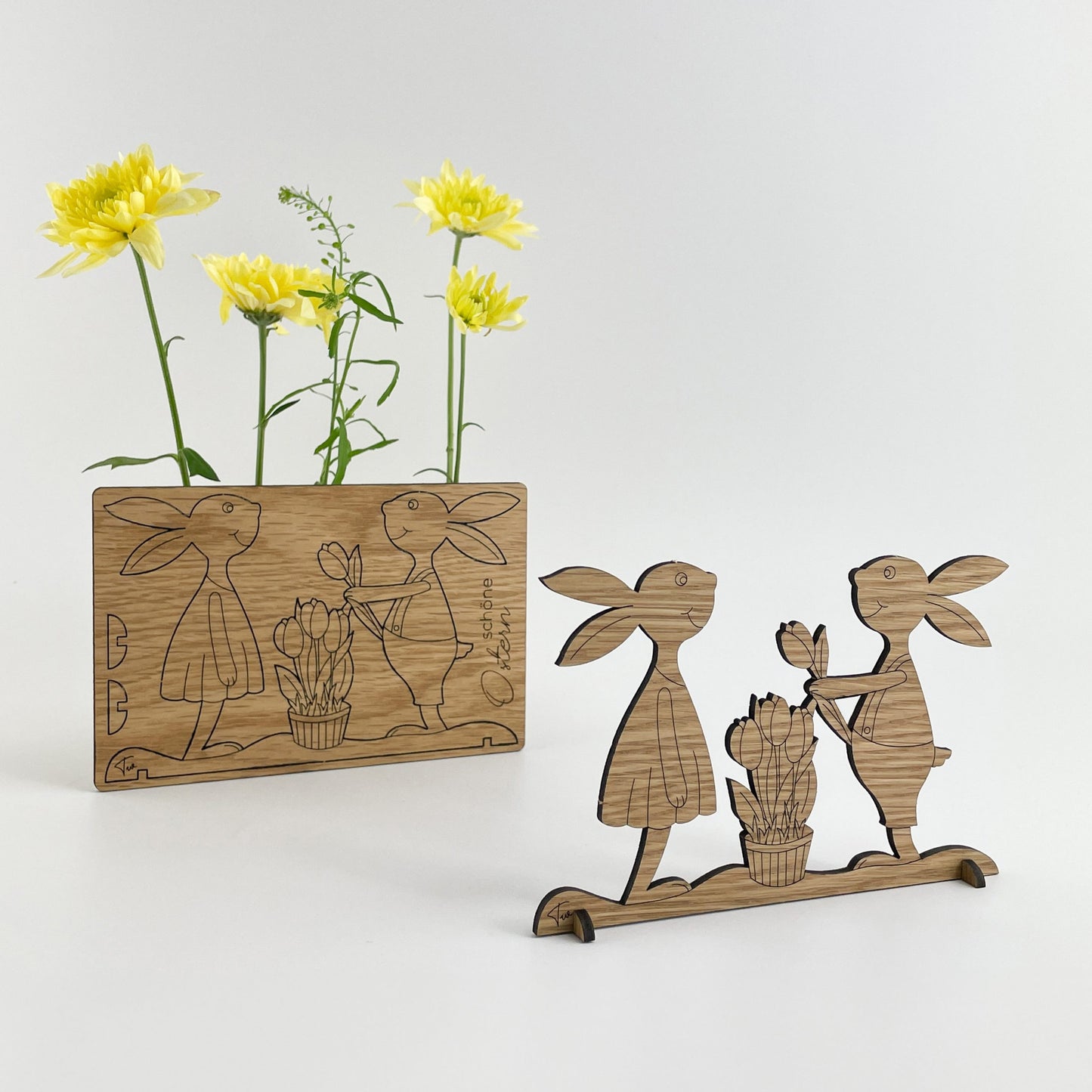 Holz-Osterkarte Papaja & Pepito mit zwei ausgeschnittenen Hasenfiguren, die auf einem Sockel stehen und sich gegenüberstehen. Im Hintergrund ist eine rechteckige Holzkarte mit einer Gravur von zwei Hasen und einem Korb zu sehen. Neben der Karte steht eine Vase mit gelben Blumen.
