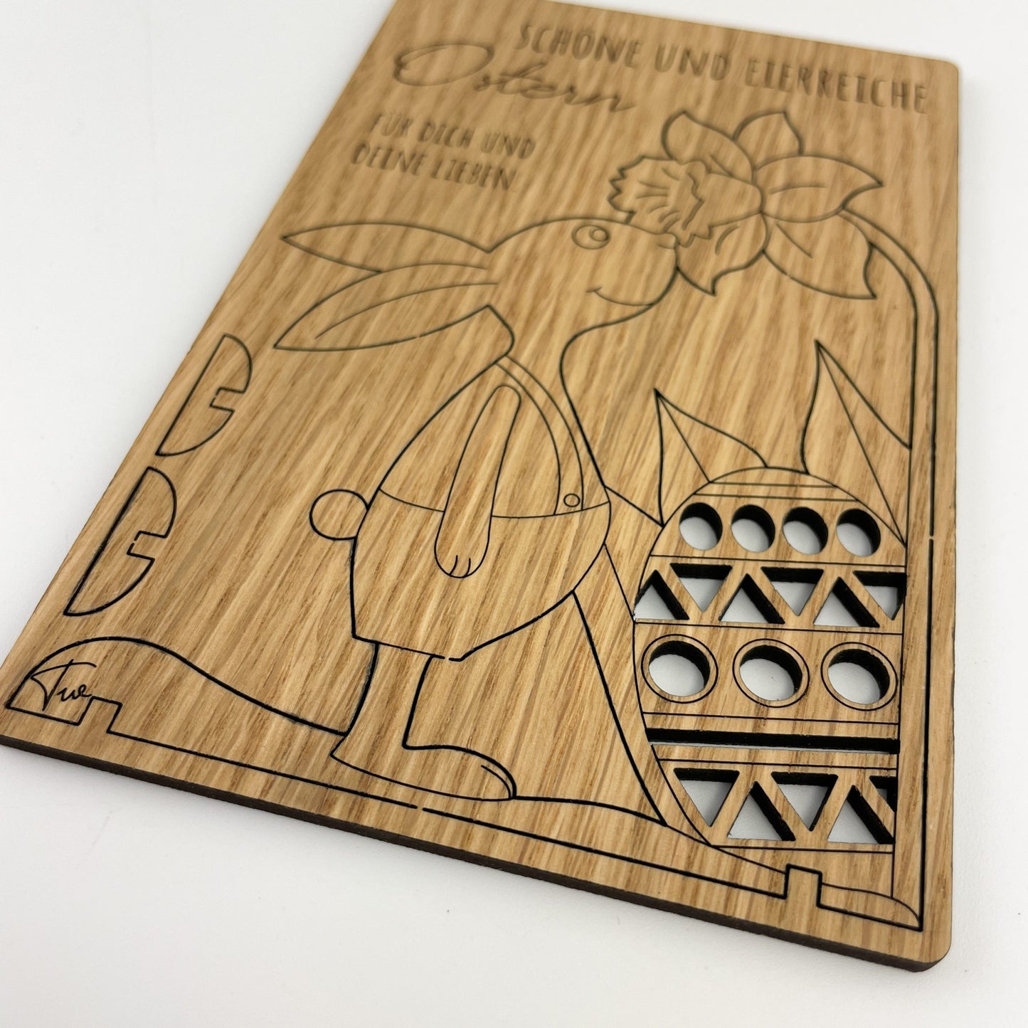 Detailansicht von Holz-Osterkarte Pino, aus Holz besticht durch ihre detailreiche Gravur und kreative Gestaltung. Darunter befindet sich eine liebevoll gestaltete Szene: Ein Hase, der ein Ei hält, ist in Bewegung abgebildet, was durch die dynamische Stellung seiner Beine angedeutet wird. Neben dem Hasen ist eine Blume zu sehen, die das Ostermotiv vervollständigt.
