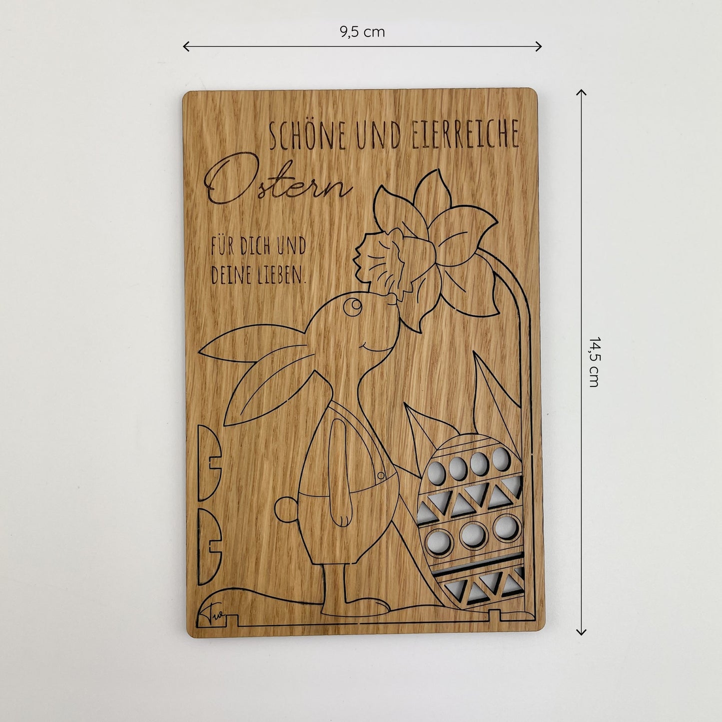 Frontalansicht von Holz-Osterkarte Pino, ist eine liebevoll gestaltete Holz-Osterkarte, die durch ihre aufwendige Laserschnitt-Technik besticht. Im oberen Teil der Karte steht in geschwungener Schrift "Schöne und eierreiche Ostern für dich und deine Lieben". Unterhalb des Textes ist ein aufrecht stehender Hase abgebildet, der ein großes Ei in der Hand hält. Neben dem Hasen befindet sich eine stilisierte Blume mit großen Blütenblättern.