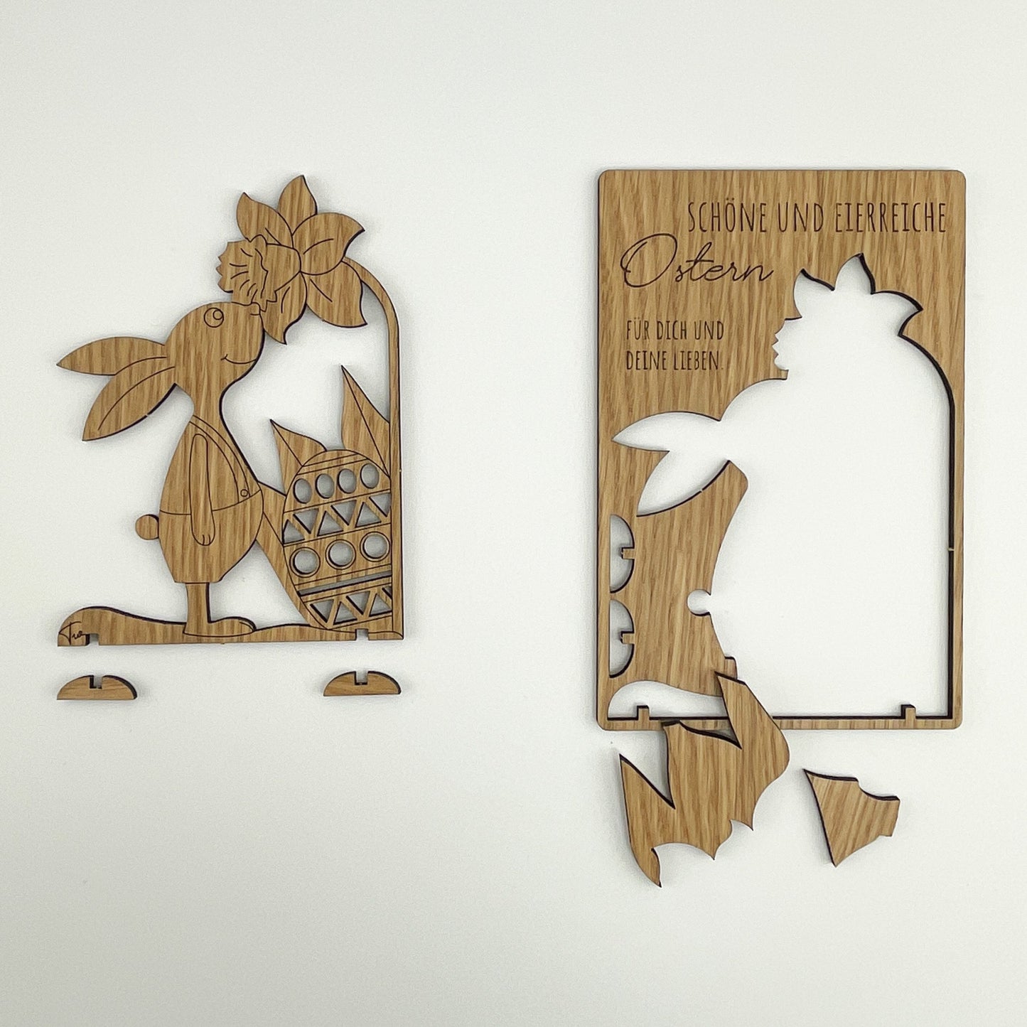 Frontalansicht von Holz-Osterkarte Pino, zeigt zwei wunderschöne Osterkarten aus Holz mit komplizierten Ausschnitten. Die linke Karte zeigt einen Hasen, der einen Korb mit Ostereiern hält, umgeben von Blumen und Blättern. Die rechte Karte enthält Text und zeigt die Silhouette eines Hasen neben einem Ei, mit Zweigen und Blättern im Hintergrund.