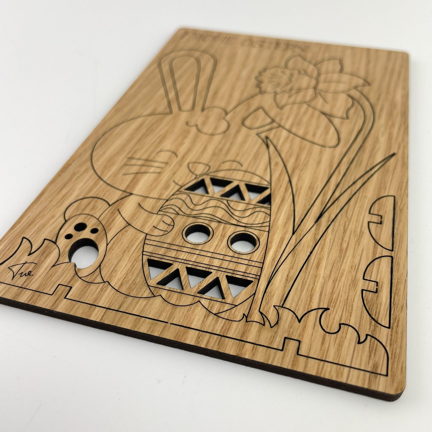 Detailansicht von Holz-Osterkarte Pascal ist ein kunstvoll gestaltetes Stück, das traditionelle Ostersymbole in einer modernen und rustikalen Ästhetik vereint. Im Mittelpunkt der Karte befindet sich ein großes Osterei, das mit verschiedenen Mustern wie Streifen und Dreiecken verziert ist. Oberhalb des Eies ist ein Hase abgebildet, der nach links schaut und dessen Ohren nach oben stehen. Links vom Osterei ist eine Tulpe zu sehen, deren Stiel sich bis zum unteren Rand der Karte erstreckt.