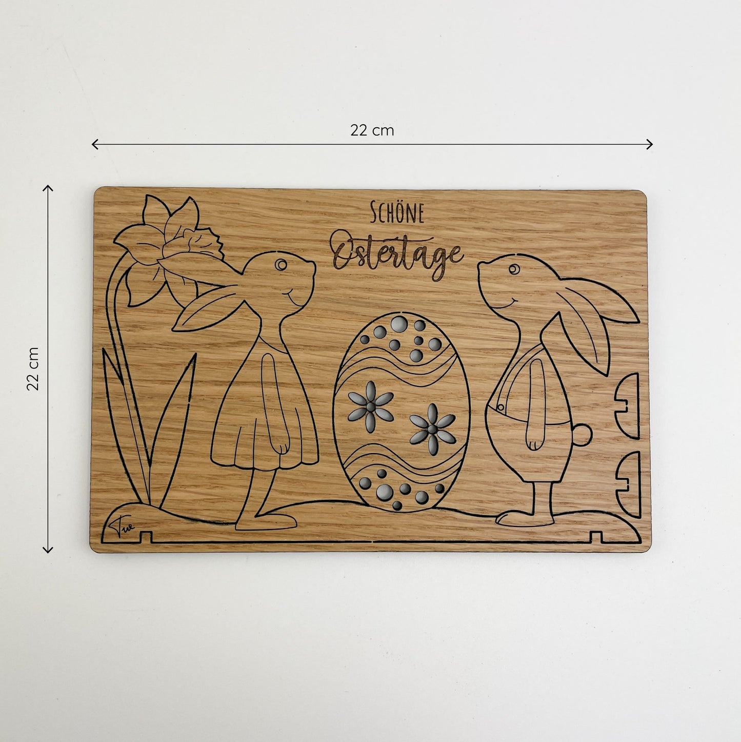 Frontansicht von Holz-Osterkarte Pippa & Pepe zeigt zwei stilisierte Hasen, einer trägt ein Kleid und der andere eine Jacke, die auf beiden Seiten eines kunstvoll gestalteten Ostereis stehen. Die Karte hat florale Muster und die Gravur ‘Schöne Ostertage’ oben. Die Abmessungen der Karte sind 22 cm x 22 cm.