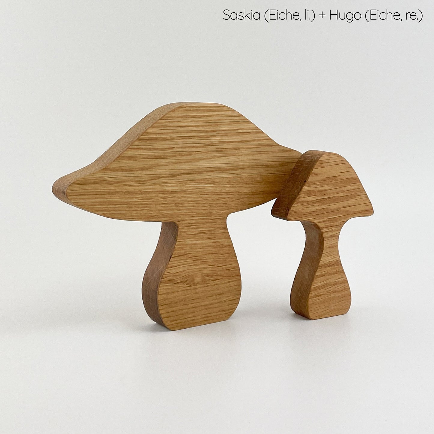 Zwei Holzpilz Deko benannt als Saskia (links) und Hugo (rechts), aus Eichenholz gefertigt, auf einem hellen Untergrund. Die größere Figur hat eine breitere Kappe und einen dickeren Stiel im Vergleich zur kleineren Figur, die eine schmalere Kappe und einen schlankeren Stiel aufweist. Beide zeichnen sich durch eine glatte Oberfläche und eine deutliche Holzmaserung aus.
