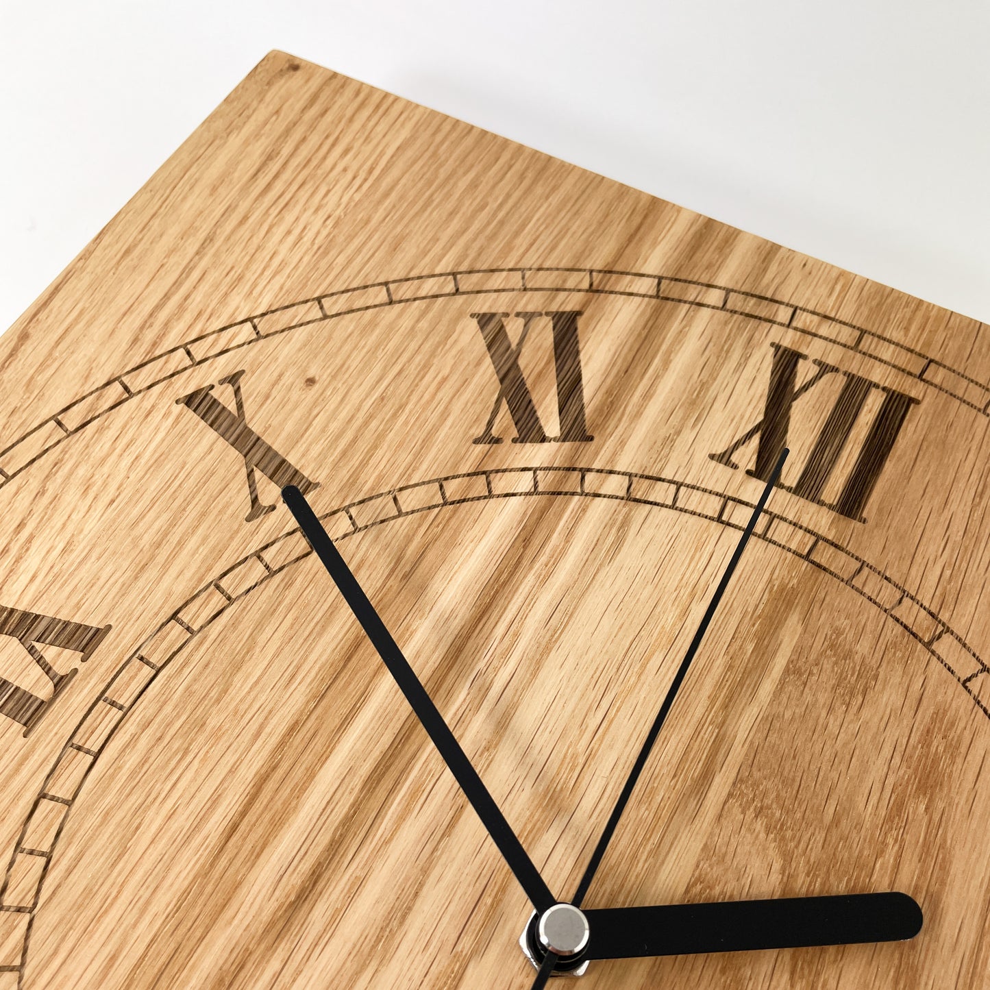 Detailansicht der quadratischen Eichenholz-Wanduhr Remus mit römischen Ziffern X, XI und XII sowie schwarzen Zeigern vor natürlicher Holzmaserung.