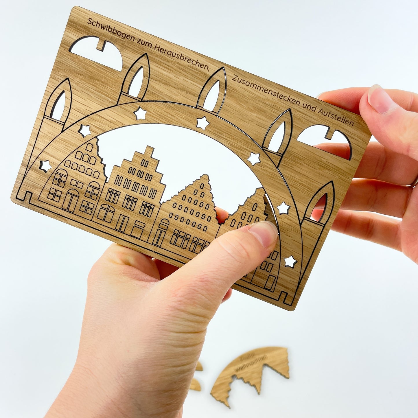 Die Weihnachtskarte aus Holz mit Schwibbogen-Motiv wird von zwei Hängen gehalten, wovon eine mit dem Daumen einen kleinen Teil des Himmels über der dargestellten Silhouette der Wismarer Altstadt herausdrückt. Die beiden Standfüße sind bereits entfernt, ebenso wie der Himmel.