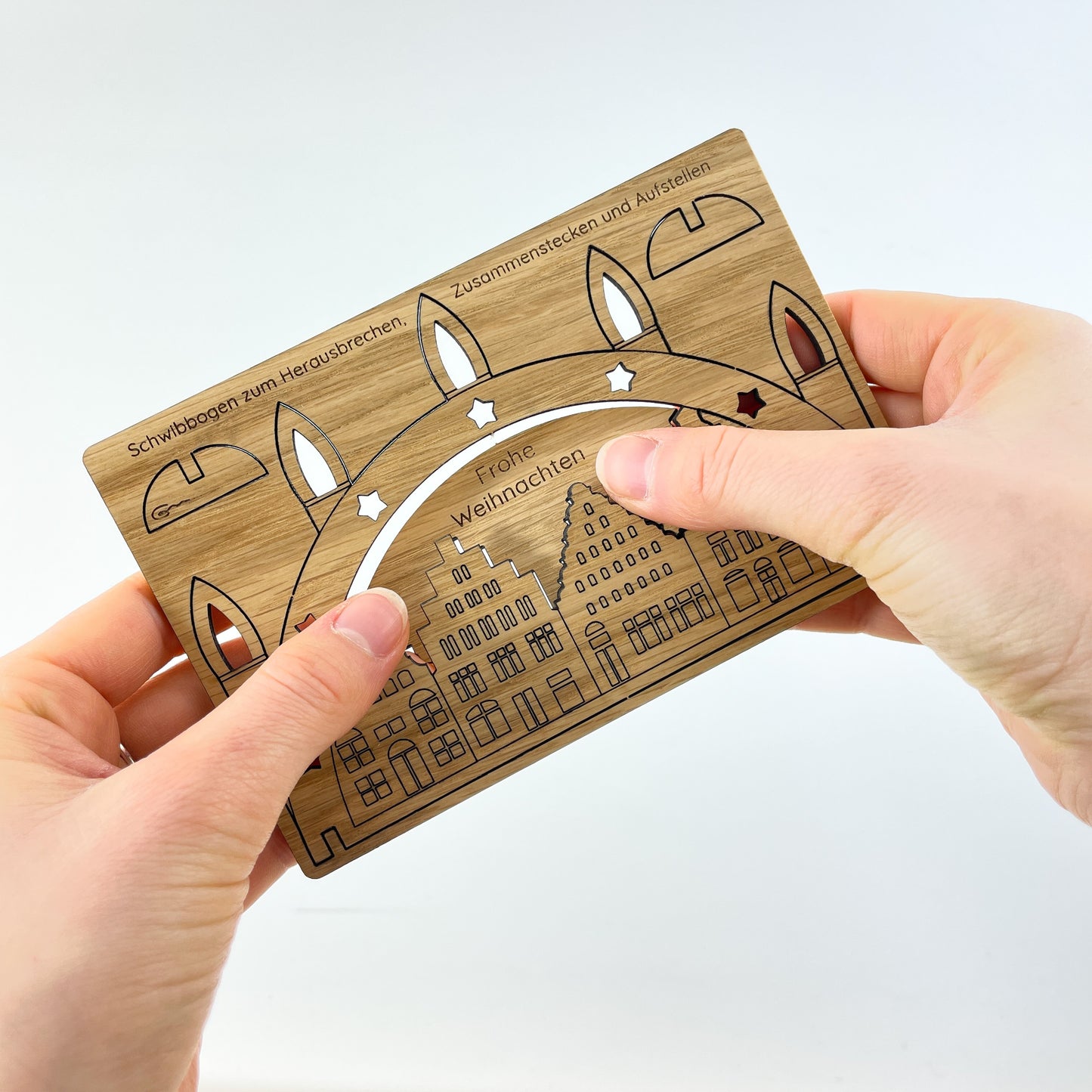 Zwei Hände halten die Weihnachtskarte aus Holz mit Wismarer-Giebelhaus-Schwibbogen-Motiv und drücken mit den Daumen einen Teil heraus, der den Himmel über den Häusern darstellt und auf dem "Frohe Weihnachten" steht.