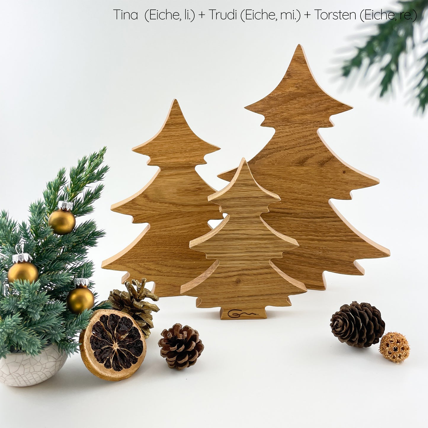 Drei unterschiedlich große Tannenbäume aus Holz stehen in der Gruppe vor einem weißen Hintergrund. Der mittlere Baum steht links hinten, der größte rechts daneben und der kleinste Tannenbaum mittig vor den beiden. Auf diesem kleinsten sieht man unten im Bereich des Stammes das Logo der Goertz Möbelmanufaktur eingebrannt. Vor und neben den drei Bäumen liegen einige Zapfen sowie eine getrocknete Orangenscheibe. Links vorn im Bild ist ein kleiner grüner Mini-Tannenbaum mit kleinen goldenen Kugeln zu sehen.