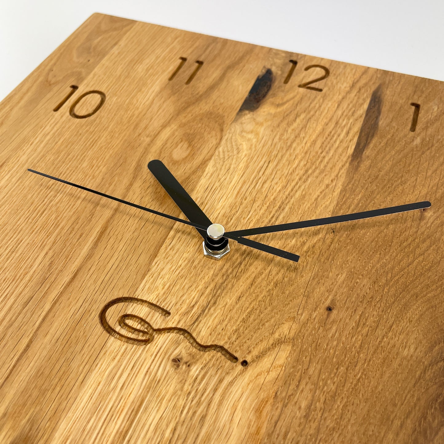 Detailansicht einer eckigen Holz-Wanduhr mit schwarzen Zeigern, bei der die Zahlen die die Stunden anzeigen eingraviert sind.