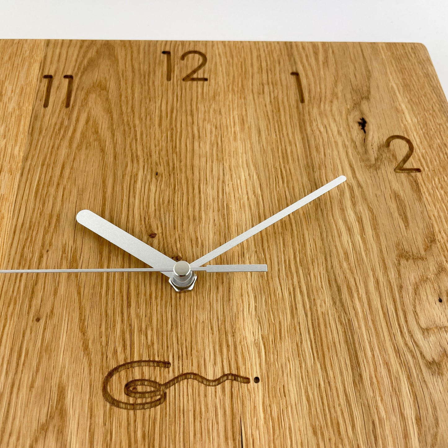 Detailansicht einer eckigen Holz-Wanduhr mit silbernen Zeigern, bei der die Zahlen die die Stunden anzeigen eingraviert sind.