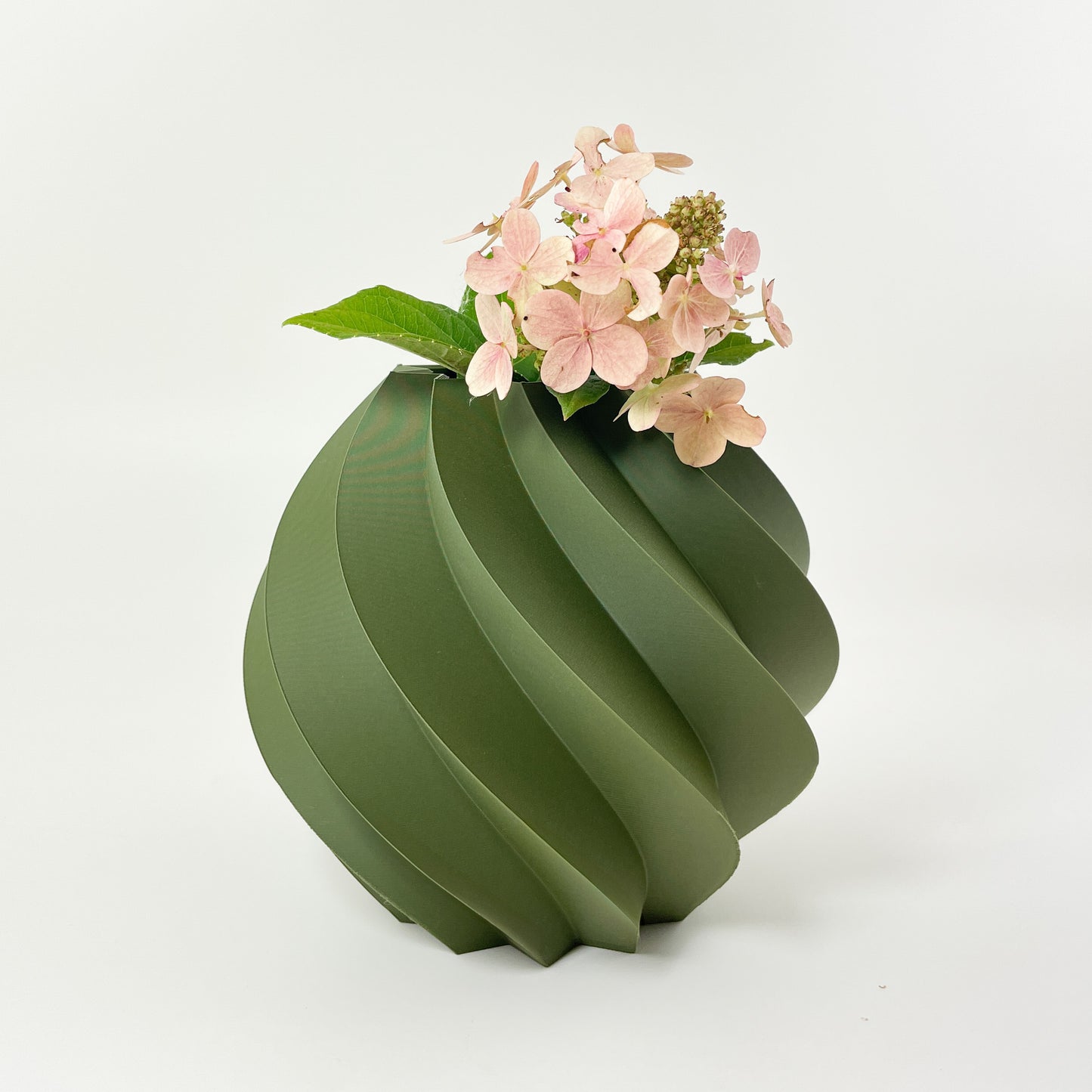 Eine runde gedrehte Vase aus mattem, moosgrünen Kunststoff vor weißem Hintergrund. Die Vase mit ihrer schönen Struktur wurde mit dem 3D-Drucker hergestellt und ist wasserdicht. Darin stehen kleine Hortensienblüten in zartem Rosa.