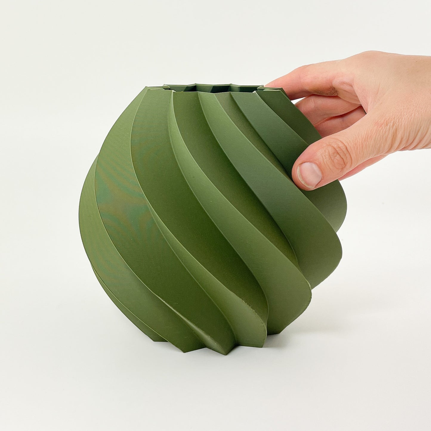 Ansicht der rundlichen olivgrünen Vase mit gedrehter Struktur aus 3D-gedrucktem Kunststoff. Rechts ragt eine Hand ins Bild, die die Vase sanft festhält. Der Hintergrund ist Weiß.