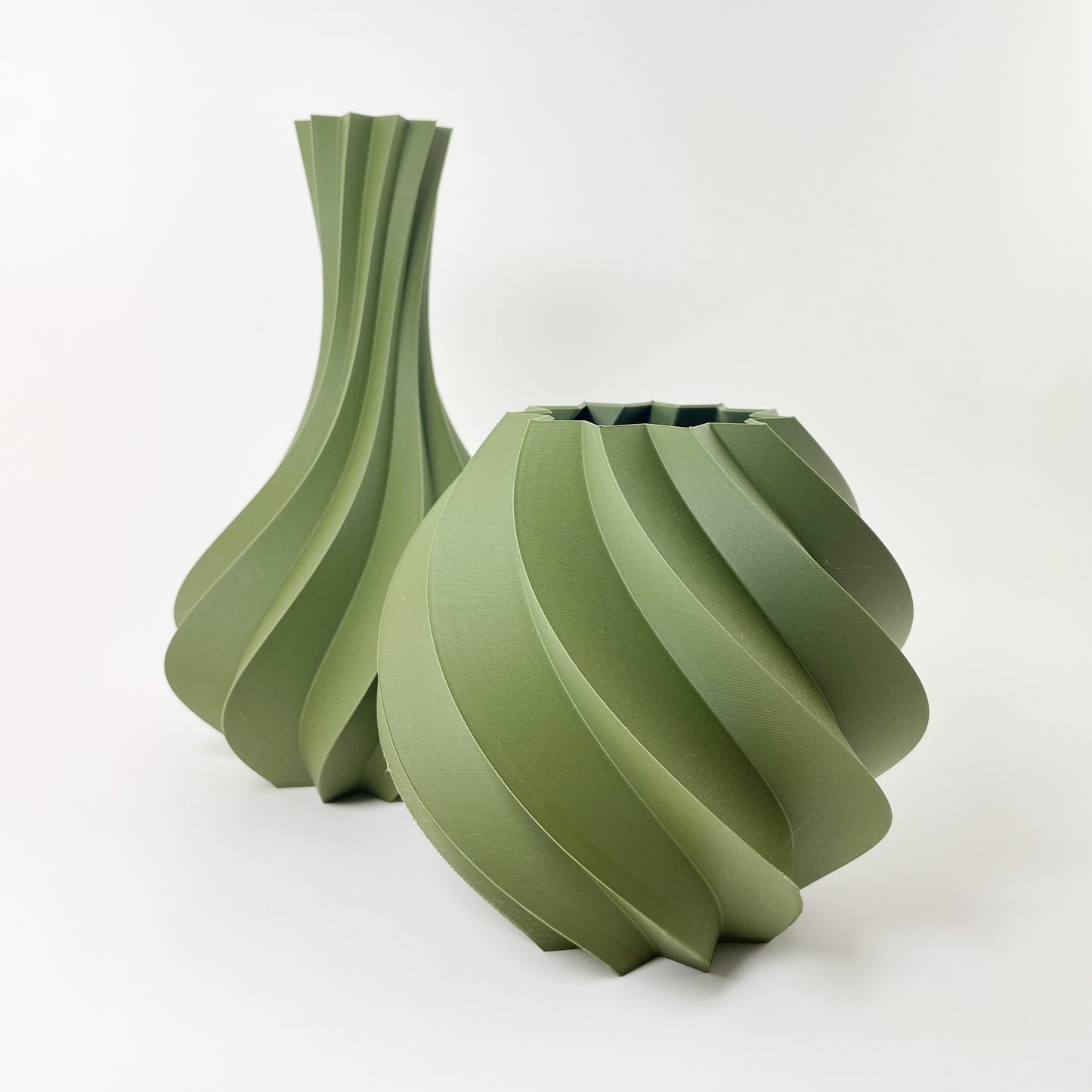 Im Vordergrund steht die rundliche 3D-gedruckte Vase aus moosgrünem Kunststoff. Leicht schräg dahinter steht links eine weitere Vase mit ähnlicher Struktur, die aber einen lang gezogenen Hals aufweist. Das Foto entstand vor einem weißen Hintergrund.
