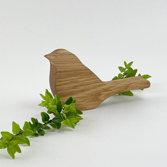 Ein Holzvogel, genannt Holzvogel Kiki, aus hochwertigem Holzmaterial, stilisiert und vereinfacht dargestellt, präsentiert auf einem weißen Hintergrund. Der Vogel zeigt glatte Konturen und keine detaillierten Merkmale. Eine grüne Pflanze mit kleinen Blättern schlängelt sich um den Schwanz des Vogels und bietet einen natürlichen Kontrast zum Holz. Dieses Bild zeigt handwerkliches Geschick und die Verwendung organischer Materialien, um Kunstwerke inspiriert von der Natur zu schaffen.