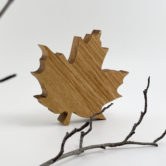 Ein Blatt aus Holz, genannt Ahornblatt Henny, mit glatter Oberfläche, das auf seiner Schnittkante als Dekoration aufgestellt wurde. Im Vordergrund einige Zweige, der Hintergrund ist Weiß.