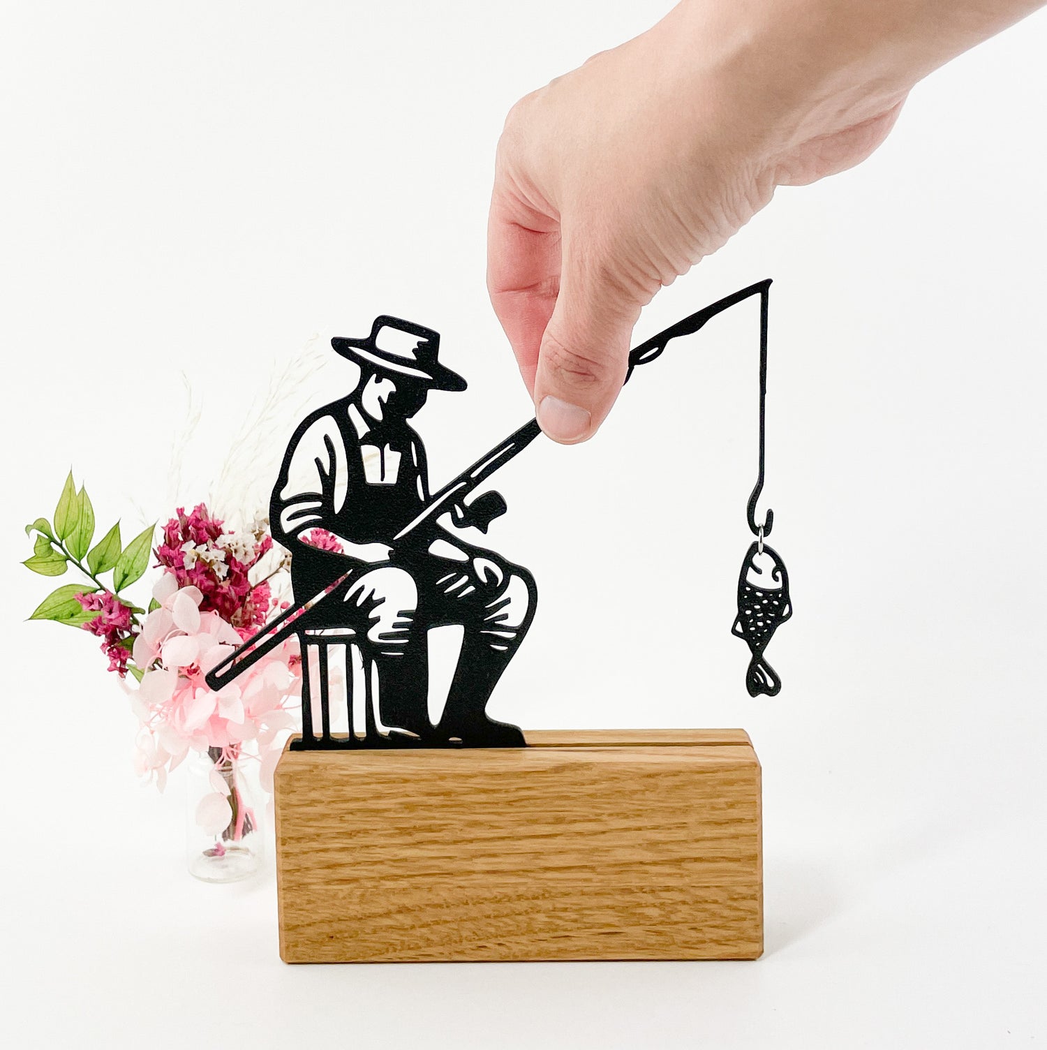 Liniensilhouette eines Anglers aus schwarzem Kunststoff, der mit dem 3D-Drucker gefertigt wurde. Er steckt in einem Halter aus massivem Eichenholz und an der Angel hängt ein kleiner Fisch, der mit einem Ring befestigt wurde. Der Angler schaut nach rechts, trägt einen Hut und ist insgesamt abstrahiert dargestellt. Eine Hand greift von oben rechts ins Bild und greift die Angel. Links hinter dem Anglermotiv steht ein kleiner Trockenblumenstrauß in rosa Farbtönen und der Hintergrund insgesamt ist Weiß.
