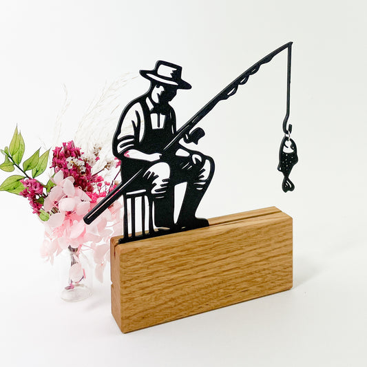 Liniensilhouette eines Anglers als Steckbild aus schwarzem Kunststoff, der mit dem 3D-Drucker gefertigt wurde. Er steckt in einem Halter aus massivem Eichenholz und an der Angel hängt ein kleiner Fisch, der mit einem Ring befestigt wurde. Der Angler schaut nach rechts, trägt einen Hut und ist insgesamt abstrahiert dargestellt. Links hinter dem Anglermotiv steht ein kleiner Trockenblumenstrauß in rosa Farbtönen und der Hintergrund insgesamt ist Weiß.