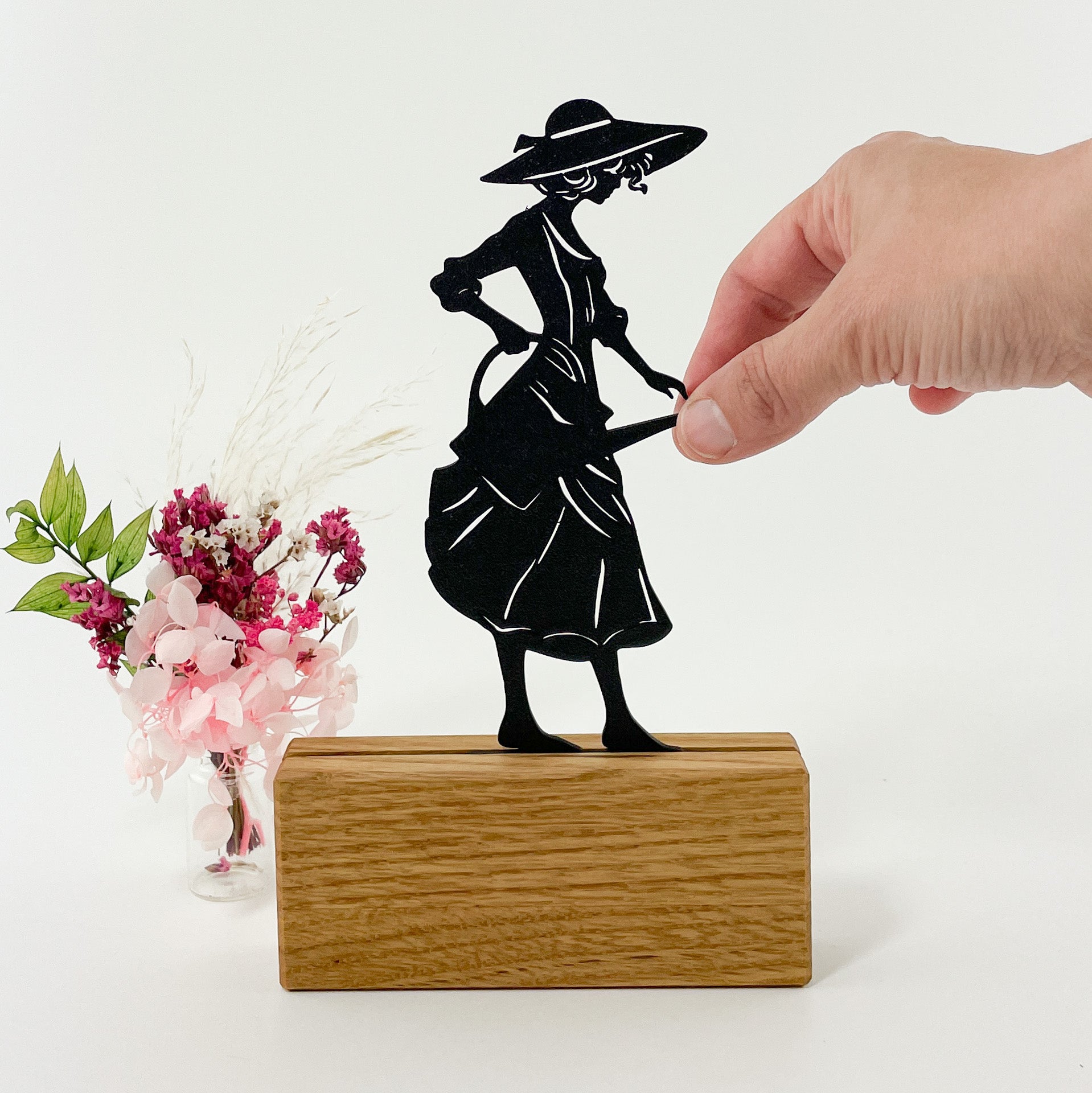 3D-gedruckte schwarze Silhouette einer eleganten Gärtnerin mit Kleid, Sonnenhut und Gießkanne, die nach rechts blickt. Sie steckt in einem Halter aus Massivholz und steht vor einem weißen Hintergrund. Rechts neben dem Kartenhalter steht ein kleiner Strauß rosafarbener Trockenblumen und es sieht so aus, als würde die Gärtnerin die Blumen gießen.