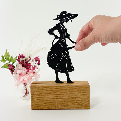3D-gedruckte schwarze Silhouette einer eleganten Gärtnerin mit Kleid, Sonnenhut und Gießkanne, die nach rechts blickt. Sie steckt in einem Halter aus Massivholz und steht vor einem weißen Hintergrund. Rechts neben dem Kartenhalter steht ein kleiner Strauß rosafarbener Trockenblumen und es sieht so aus, als würde die Gärtnerin die Blumen gießen.