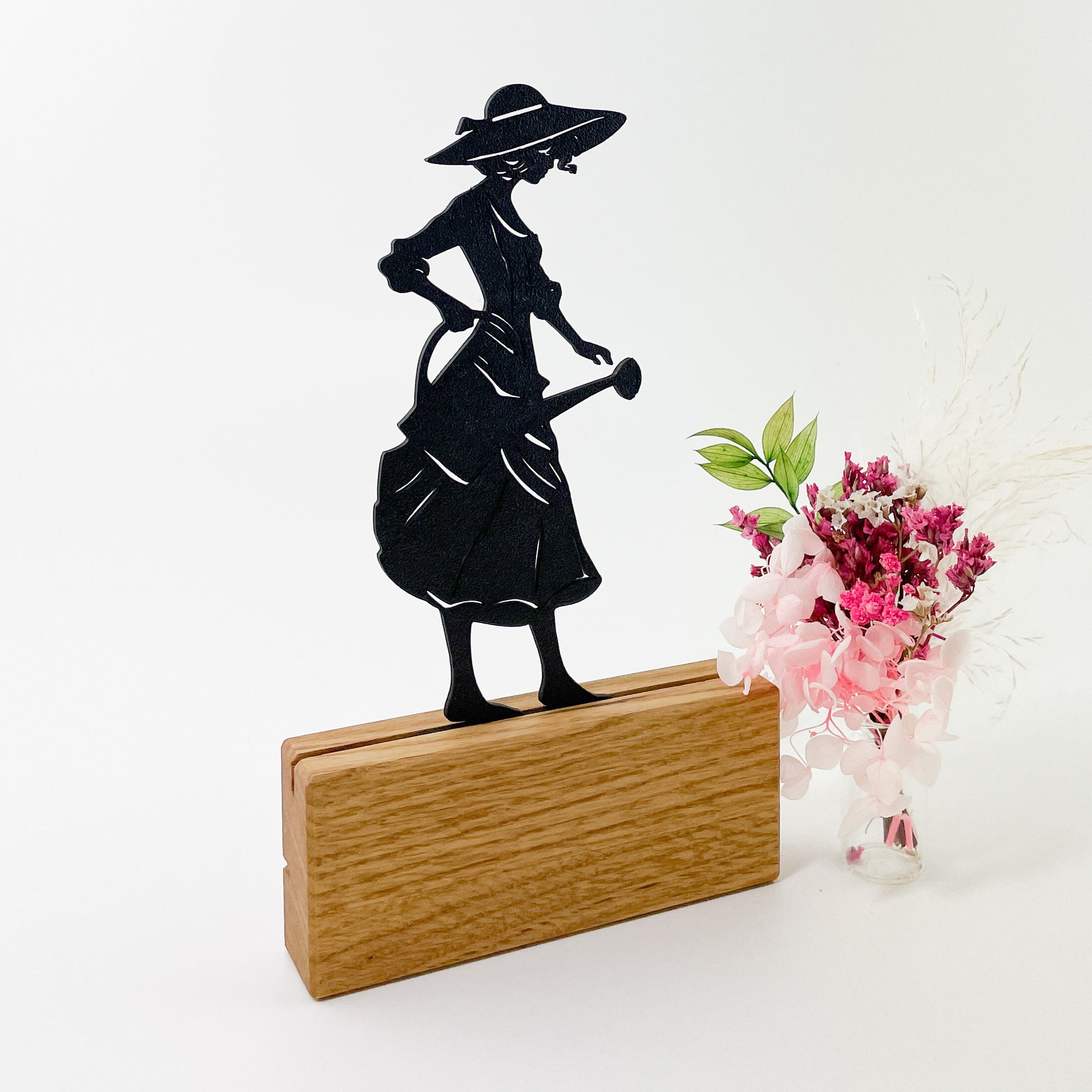 3D-gedruckte schwarze Silhouette einer eleganten Gärtnerin mit Kleid, Sonnenhut und Gießkanne, die nach rechts blickt. Das Steckbild steckt in einem Halter aus Massivholz und steht vor einem weißen Hintergrund. Rechts neben dem Kartenhalter steht ein kleiner Strauß rosafarbener Trockenblumen und es sieht so aus, als würde die Gärtnerin die Blumen gießen.