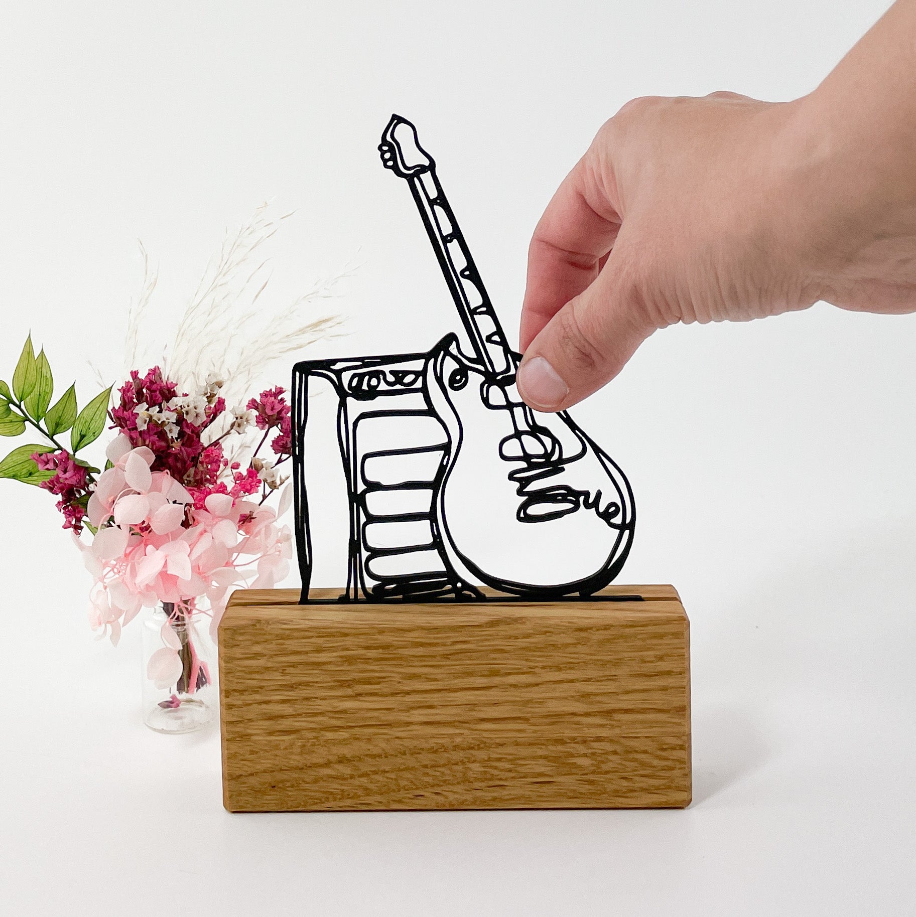 3D-Druck einer E-Gitarre mit Verstärker im Stil einer Linienzeichnung aus schwarzem Kunststoff. Das Steckbild steht in einem Halter aus massivem Eichenholz vor weißem Hintergrund. Rechts ragt eine Hand ins Bild, die mit zwei Fingern die Gitarre anfasst und links hinter dem Kartenhalter steht ein kleiner rosafarbener Trockenblumenstrauß.
