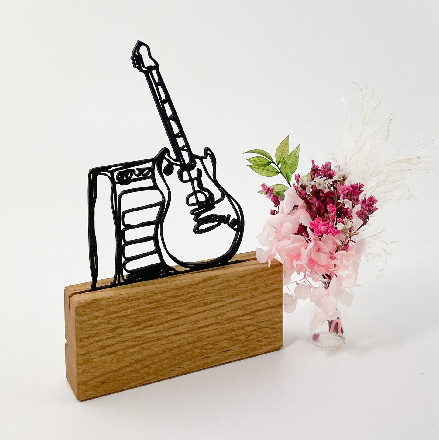 3D-gedruckte Linienzeichnung einer E-Gitarre, die an einem Verstärker lehnt. Das Steckmotiv ist aus schwarzem Kunststoff gefertigt und in einem Halter aus Massivholz platziert. Rechts daneben steht ein kleiner Trockenblumenstrauß in rosa Farbtönen und der Hintergrund ist weiß.