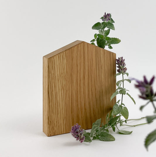 Ein Holz Dekohaus steht auf einer schlichten Oberfläche, an den Zweige von grünen und violett blühenden Pflanzen lehnen. Der Kontrast zwischen den natürlichen Elementen und dem glatten, bearbeiteten Holz schafft eine visuell harmonische Szene.